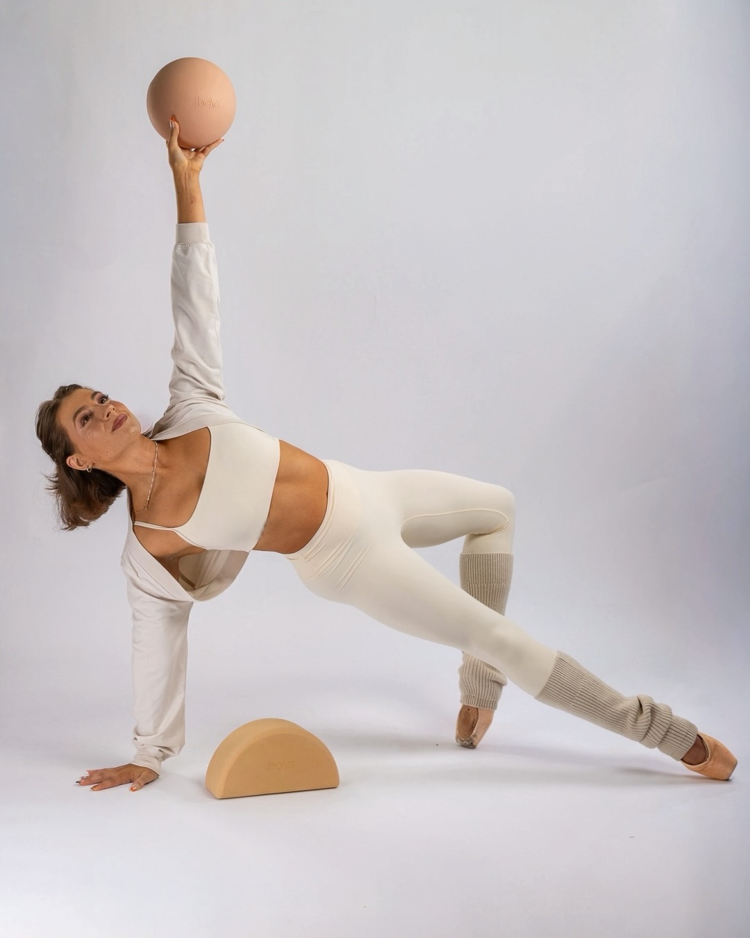 Woman in white athletic wear balances a ball on her raised hand while supporting herself with one arm and foot on the ground.