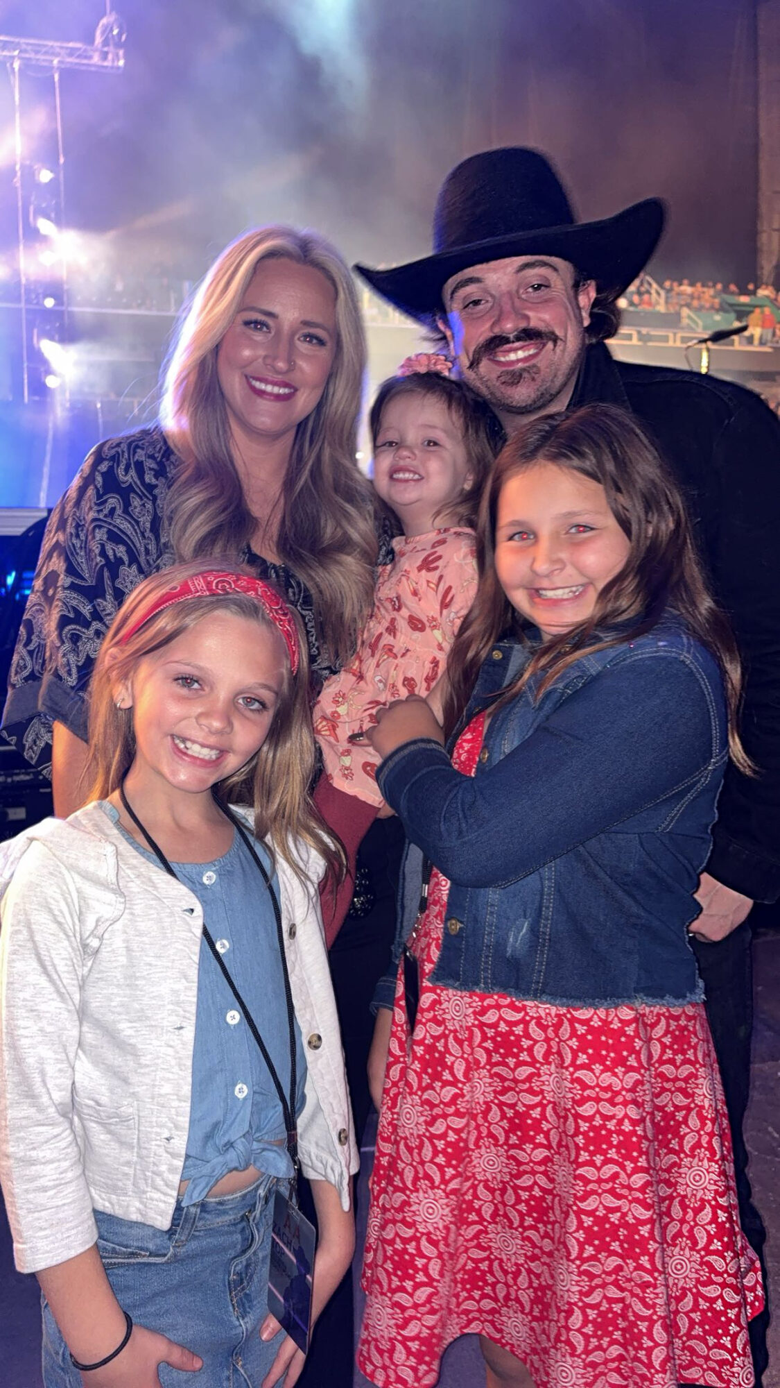Group of five people, including a man in a cowboy hat, smiling, with two young girls and two women, in a festive setting.