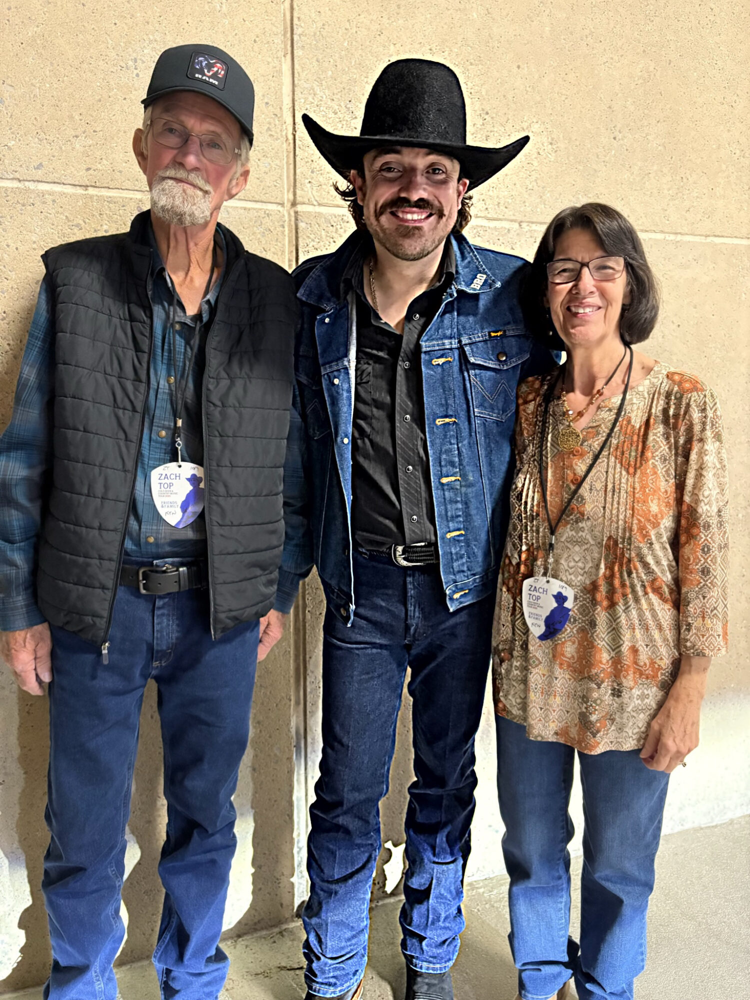 Three people standing together, smiling, against a beige wall. The person in the middle wears a black hat and denim jacket.