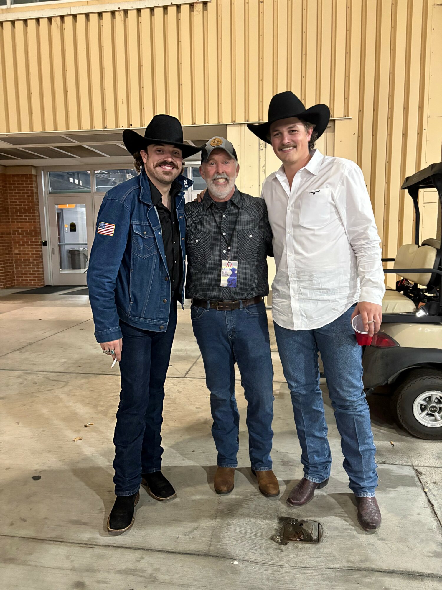 Three men wearing cowboy hats and casual clothing standing outdoors in front of a building and a golf cart.