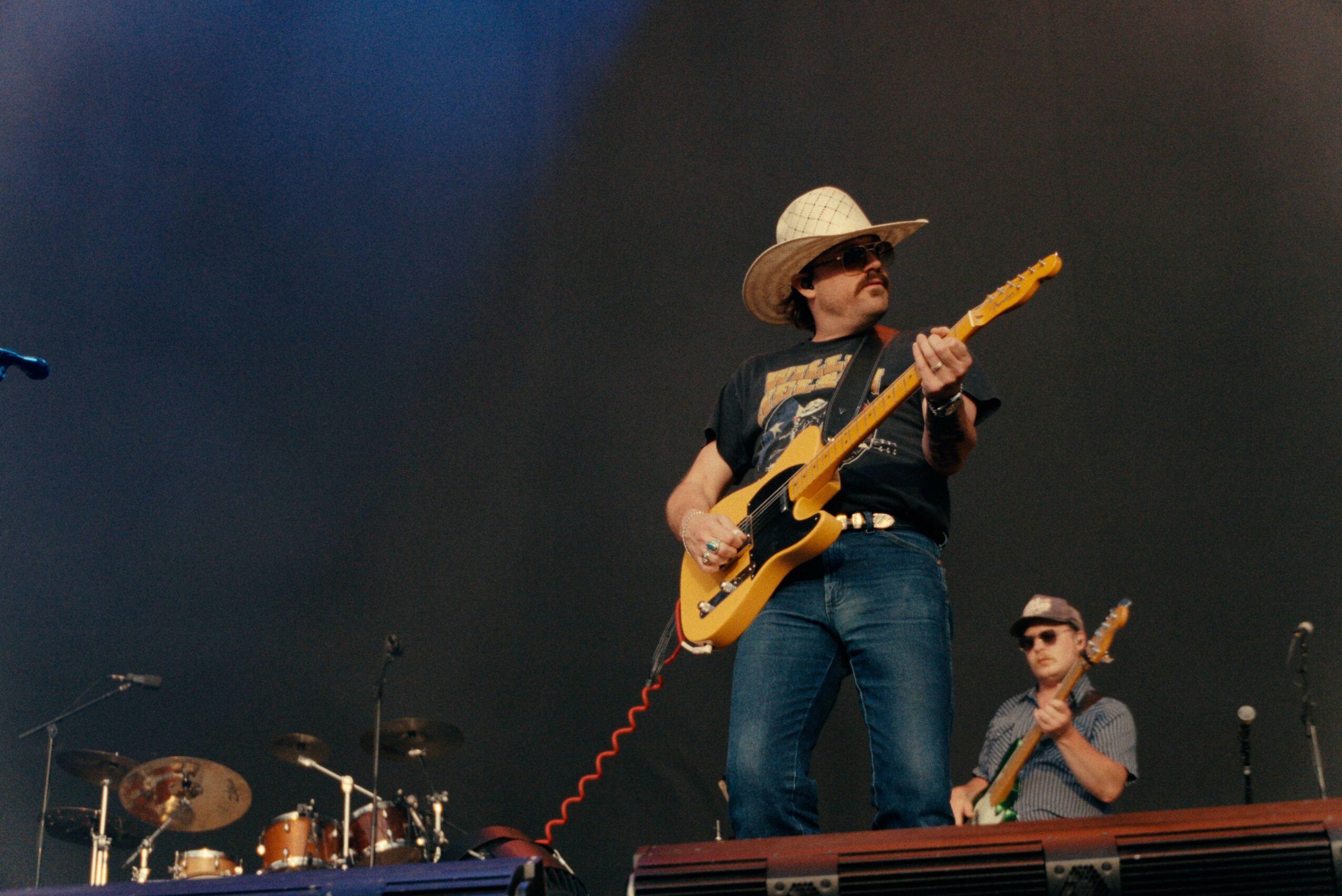 Person wearing a straw hat and sunglasses playing an electric guitar on stage with a drummer in the background.