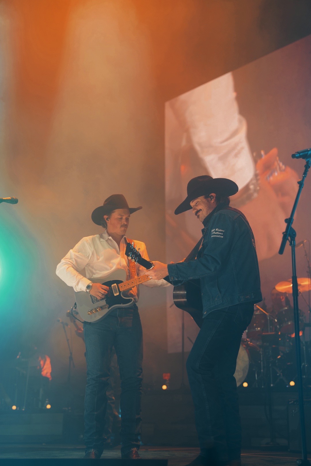 Two musicians on stage, one playing guitar and the other dancing, with colorful stage lighting and a large screen in background.