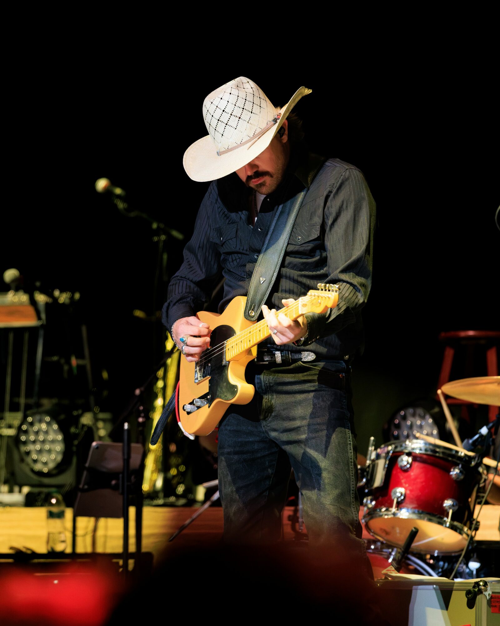 Person wearing a large hat playing an acoustic guitar on stage with drums and equipment in background.