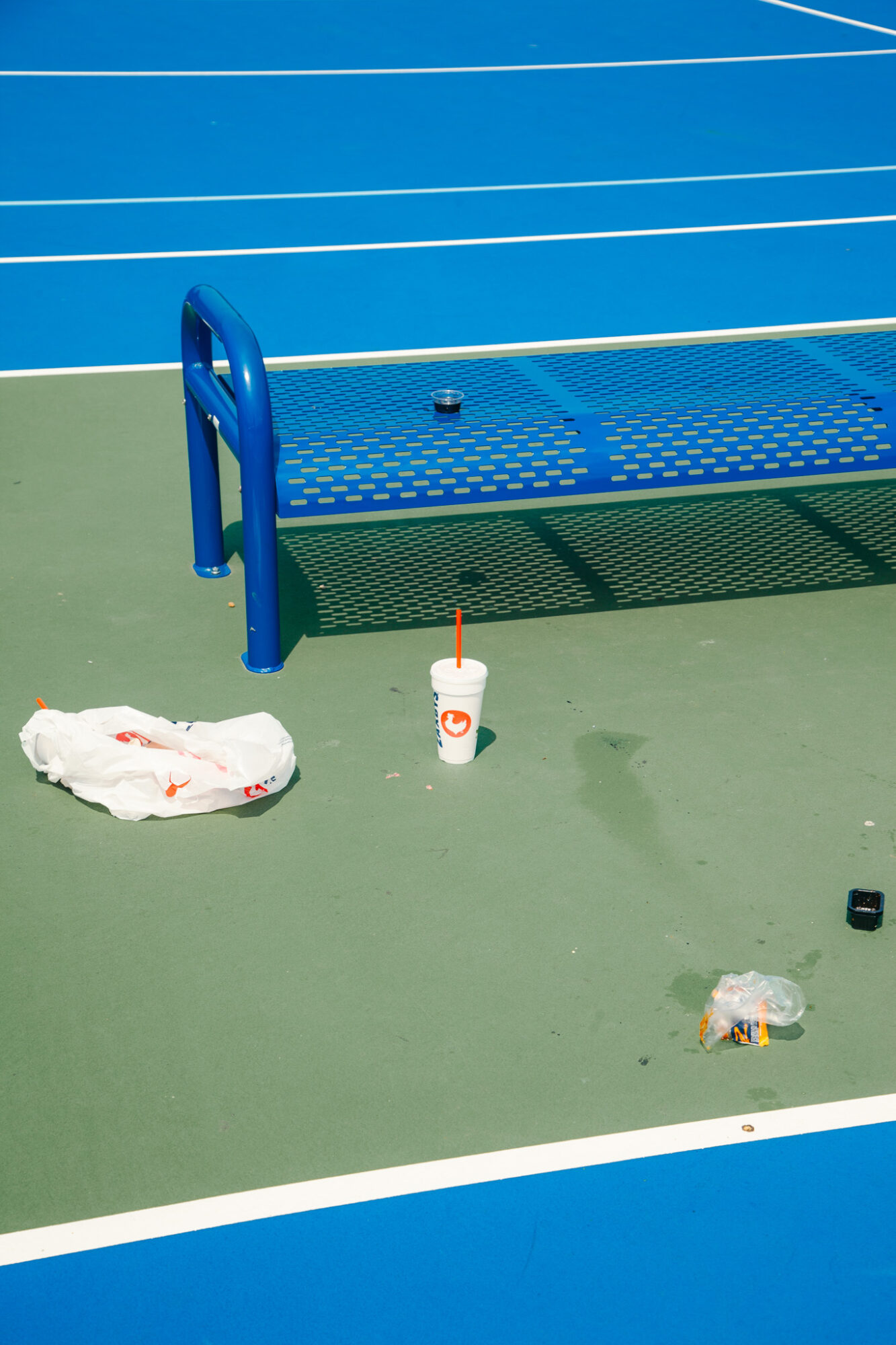 Blue bench on a green and blue sports court with a towel, drink, and small objects nearby.