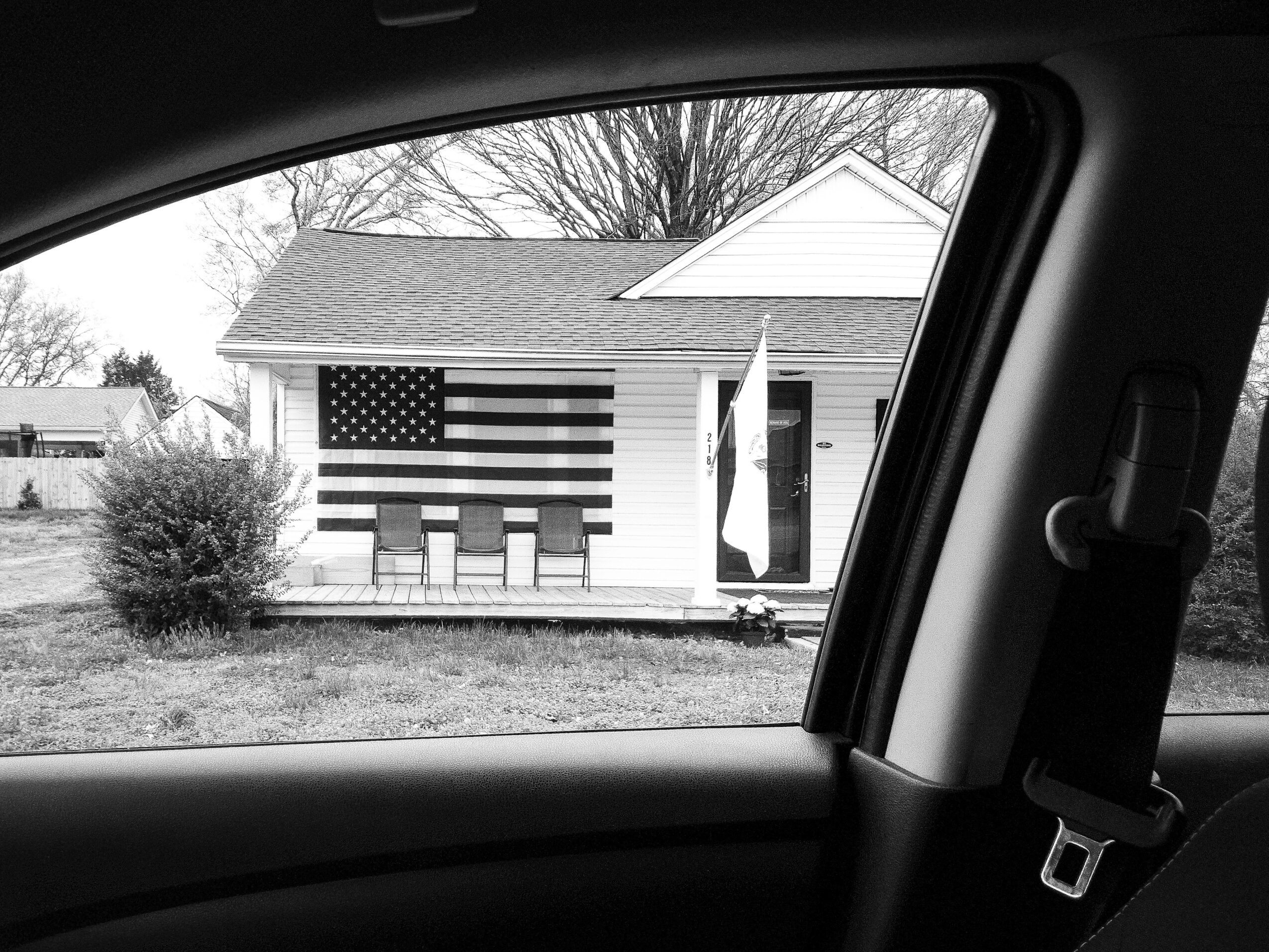 View through a car window showing a house with an American flag and a bush outside.