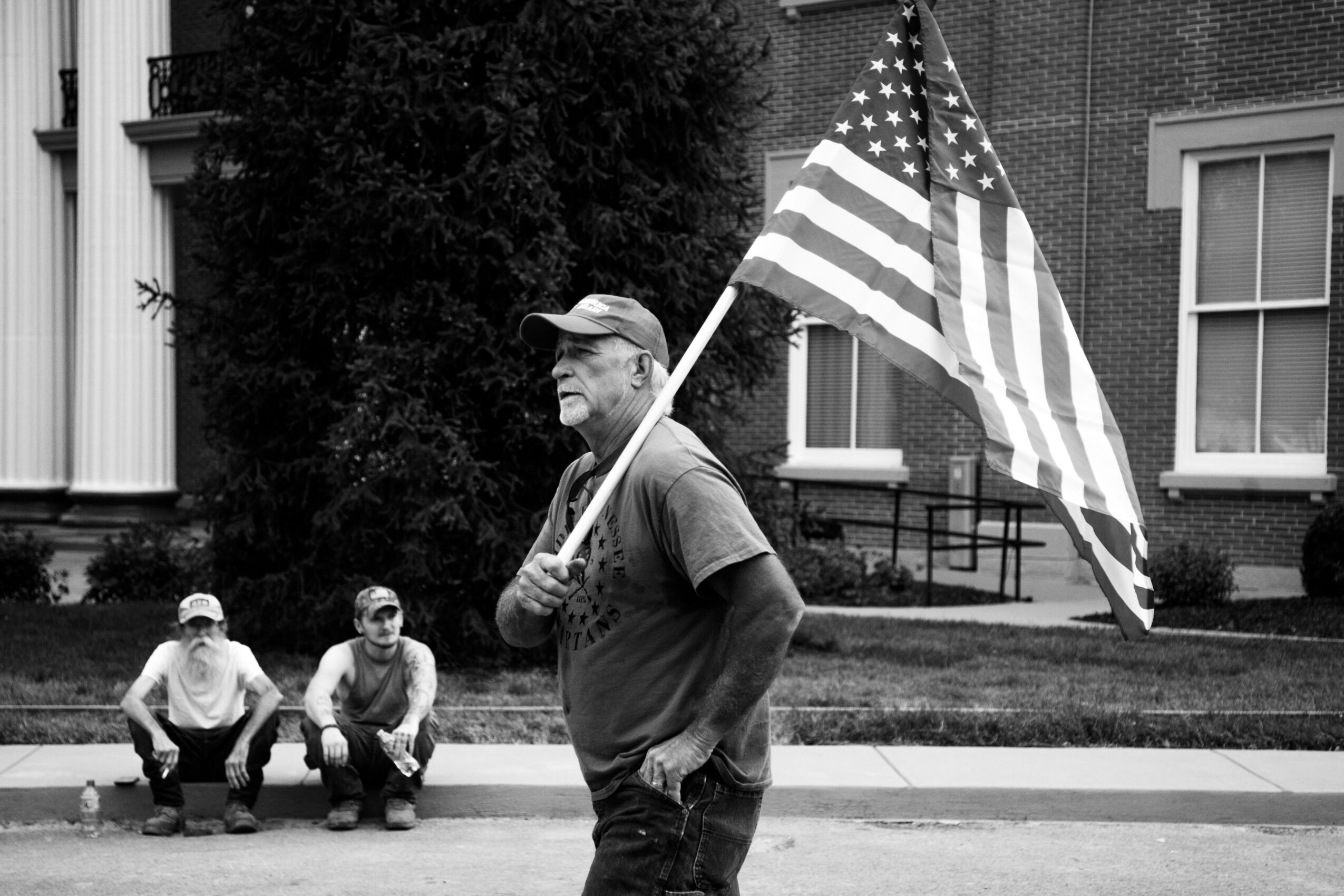 Older man holding American flag, standing outdoors with two children sitting on the sidewalk behind him, buildings in background.