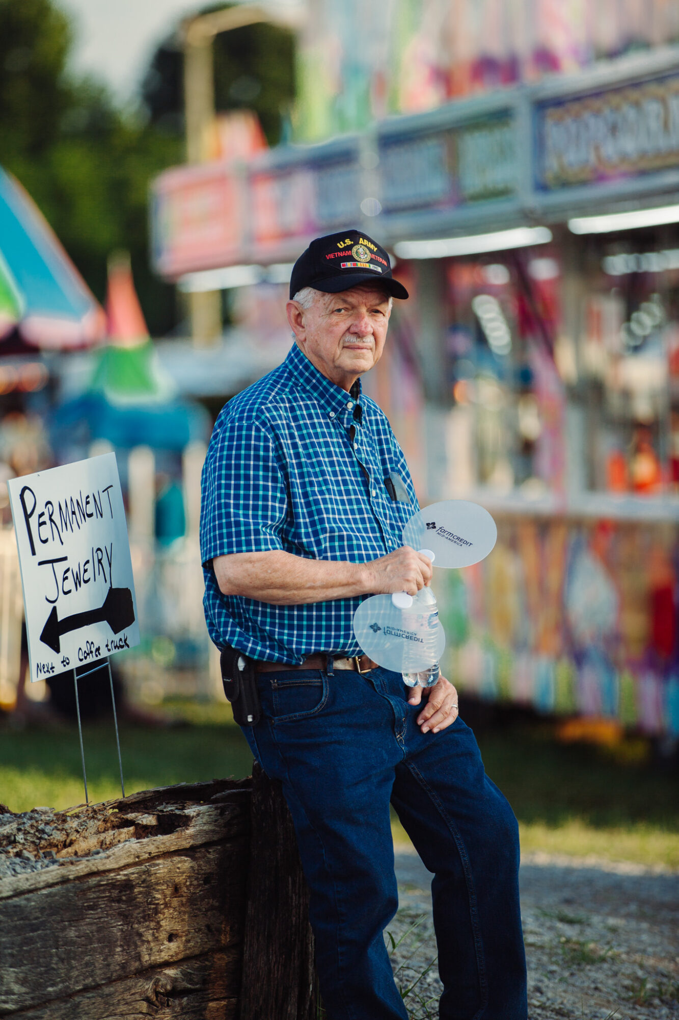 Man in blue checkered shirt and cap holding balloons at outdoor fair, colorful rides in background.