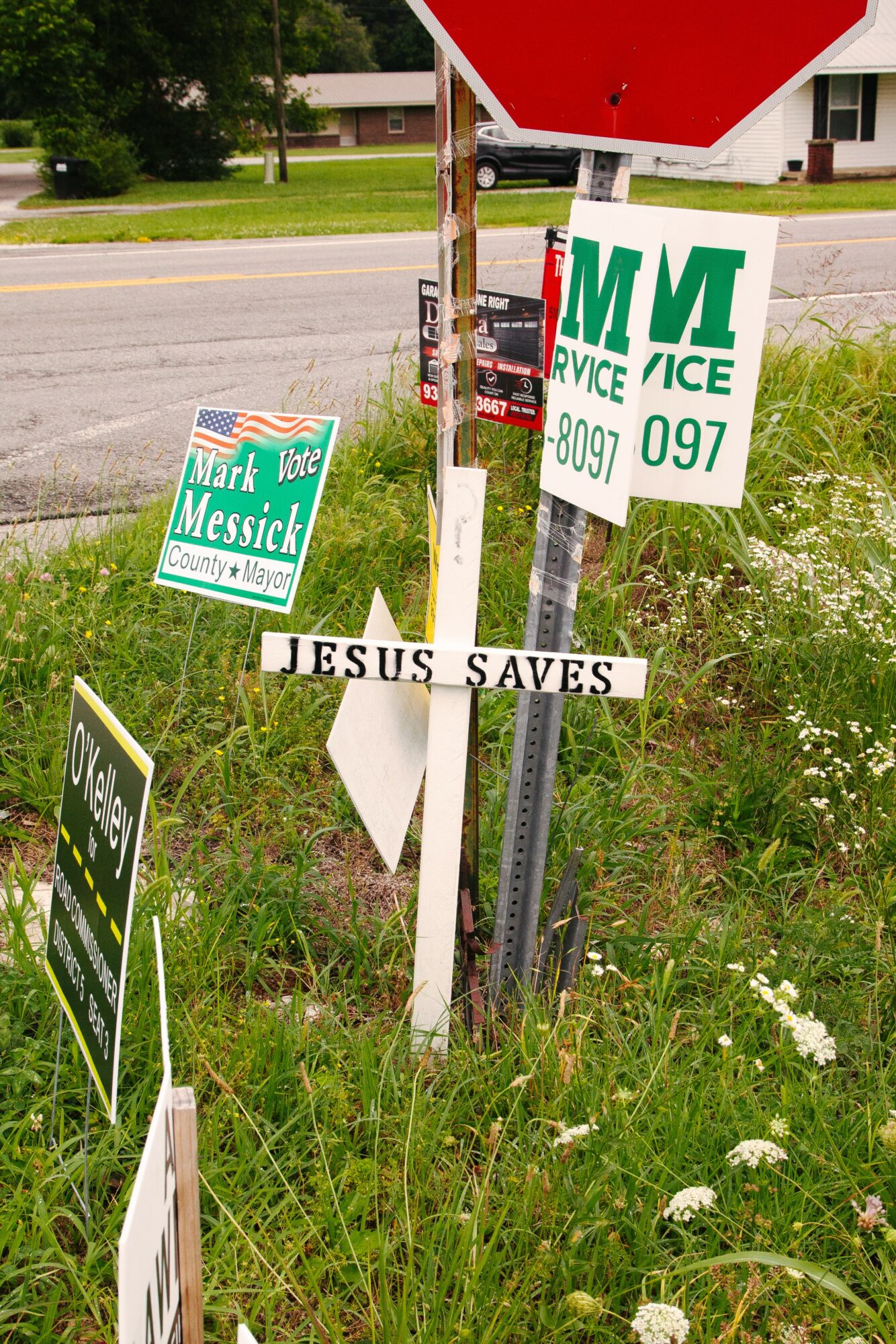 Multiple signs including political signs, a cross with the message 'JESUS SAVES,' and a stop sign, in a grassy area.