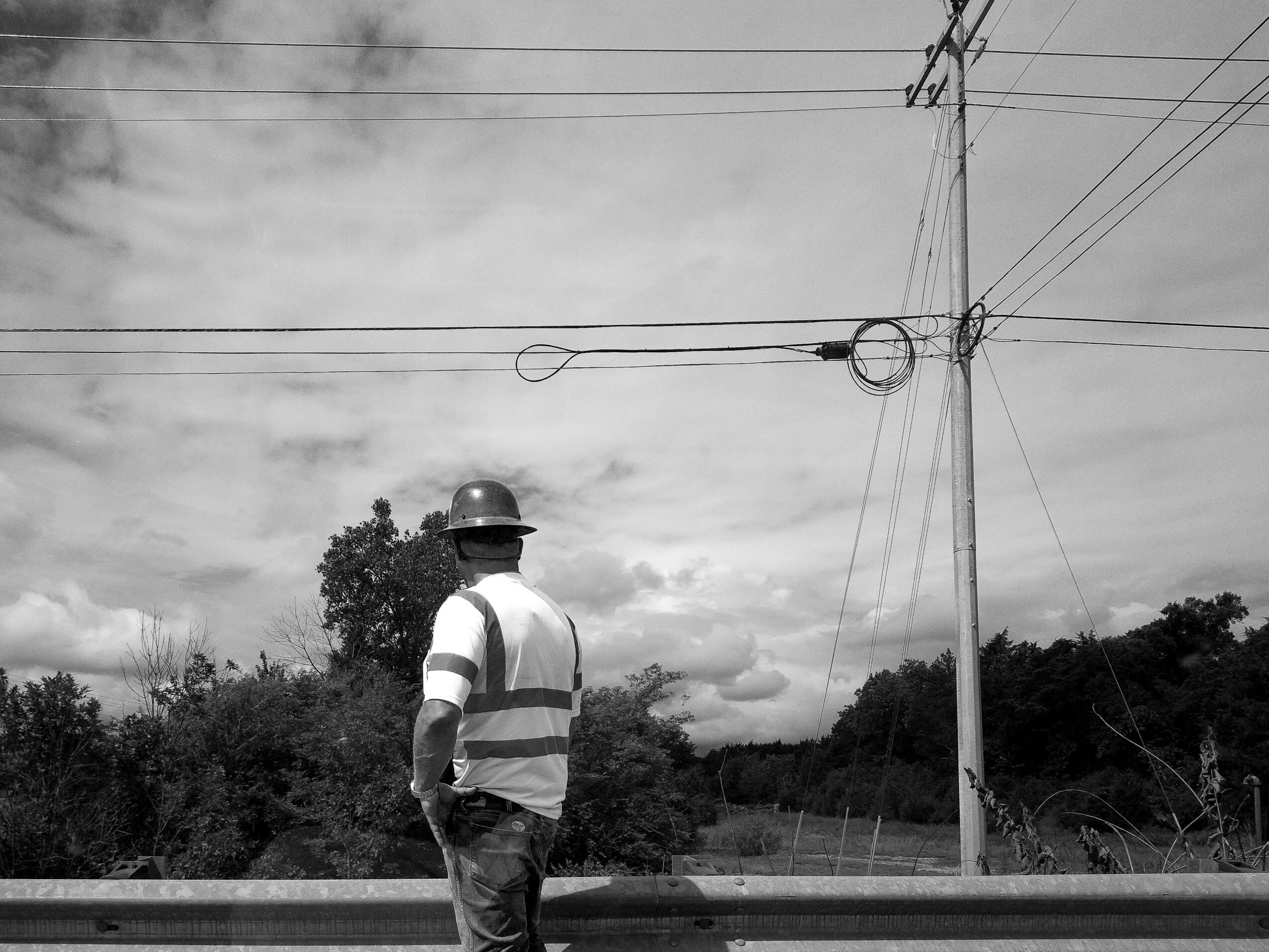 Person wearing a helmet and striped shirt standing outdoors near power lines and trees.
