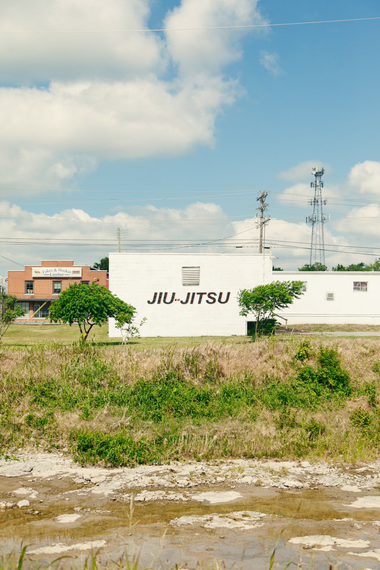 White building with black text 'JIU-JITSU' on it, green grass, trees, and a blue sky with clouds.