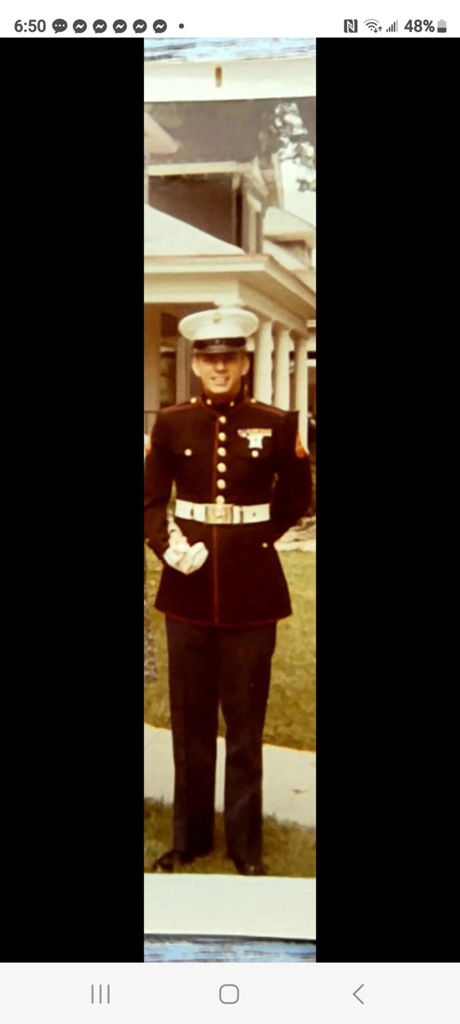 Person in military uniform standing outdoors in front of a building, wearing a hat and sunglasses.
