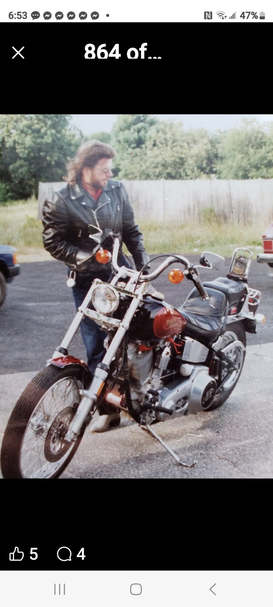 Person with long hair and leather jacket standing next to a motorcycle outdoors, trees and a fence in background.