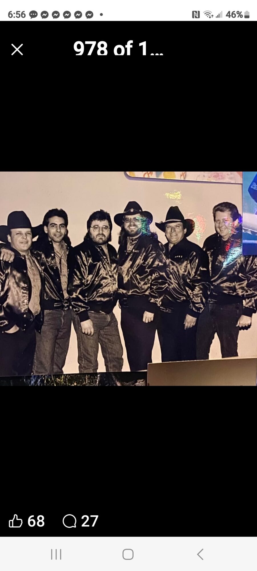 Group of six men wearing matching shiny jackets and hats, standing indoors against a plain background.