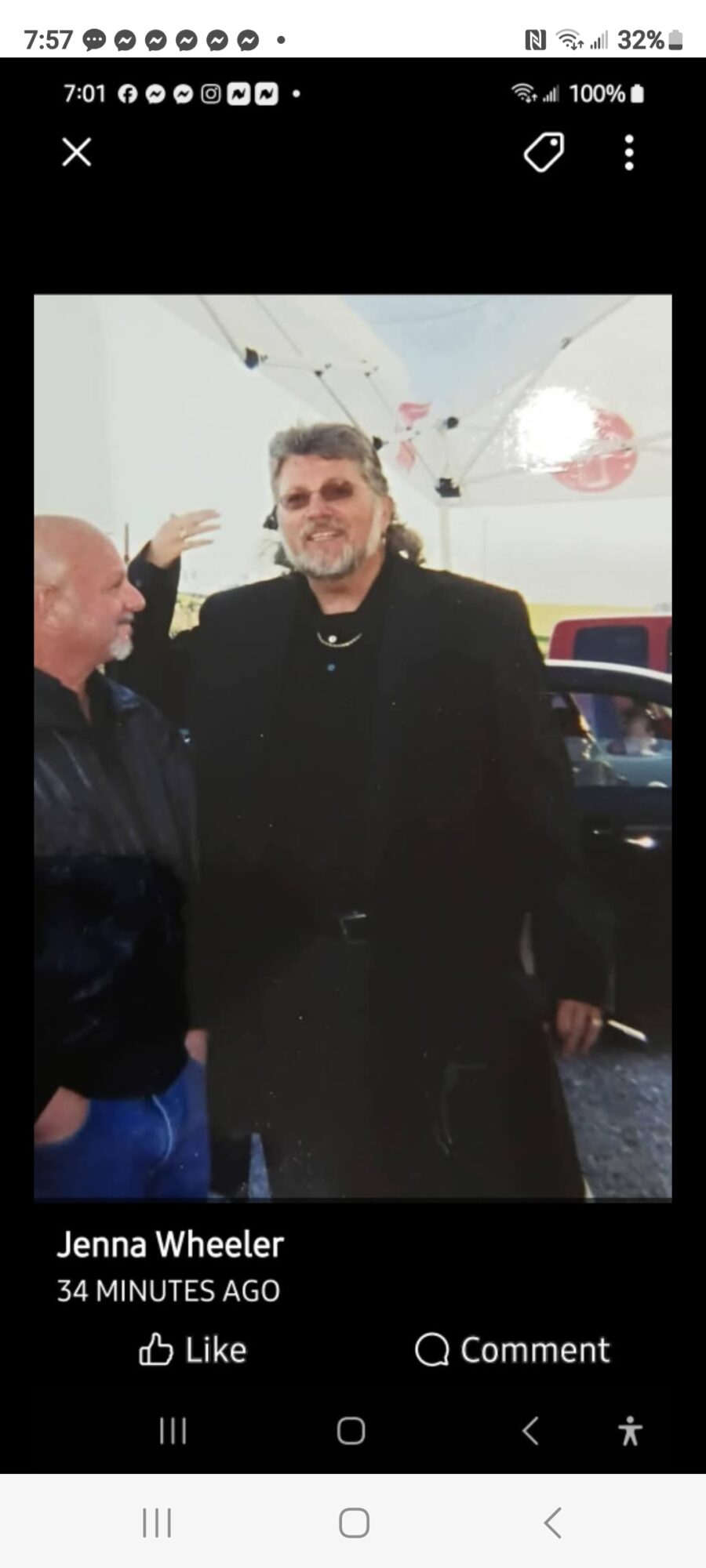 Two men standing outdoors, one with gray hair and beard, the other bald, smiling and talking, with a car in the background.