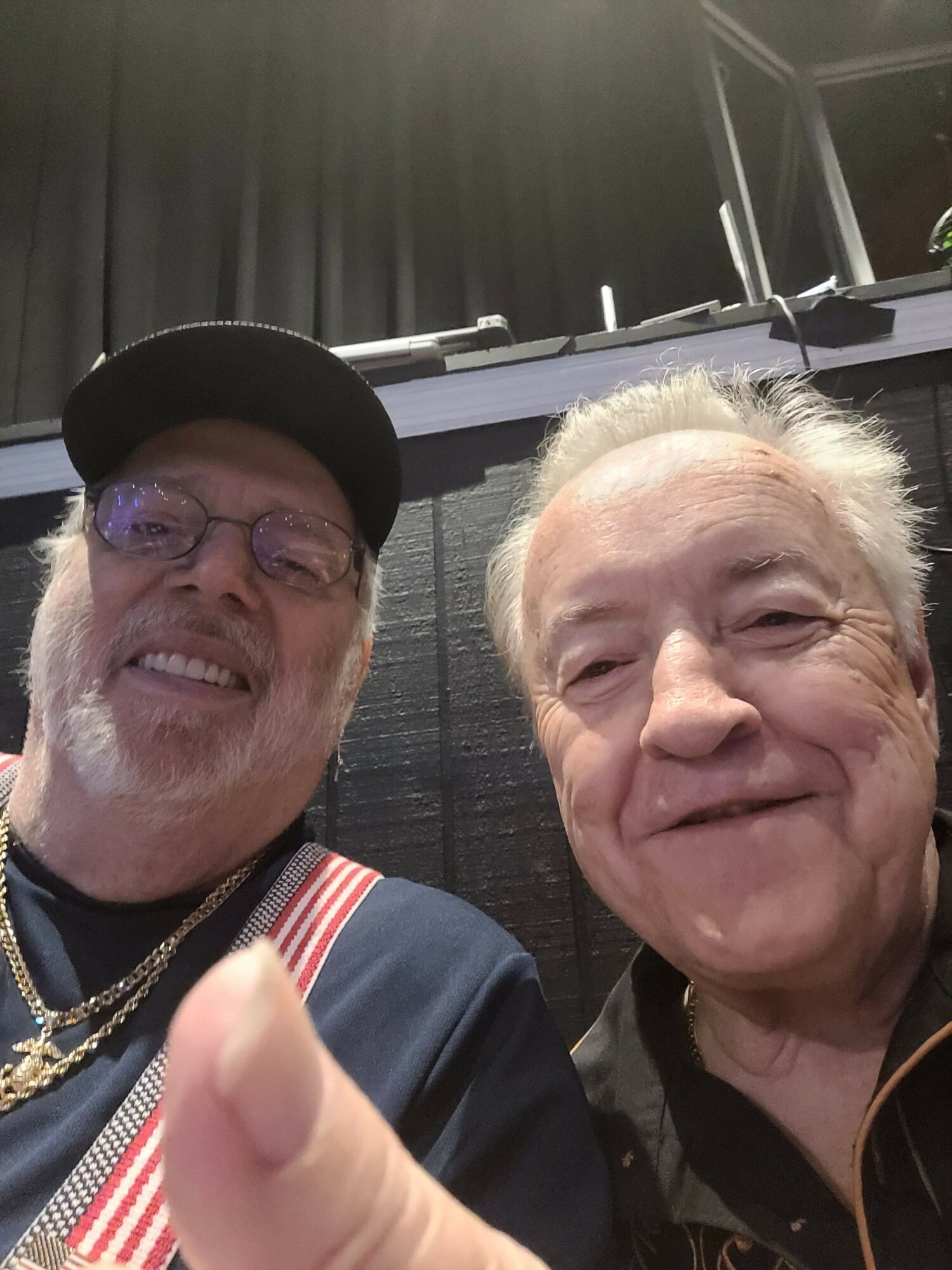 Two men smiling and posing for a photo, one wearing glasses and a cap, the other with white hair.