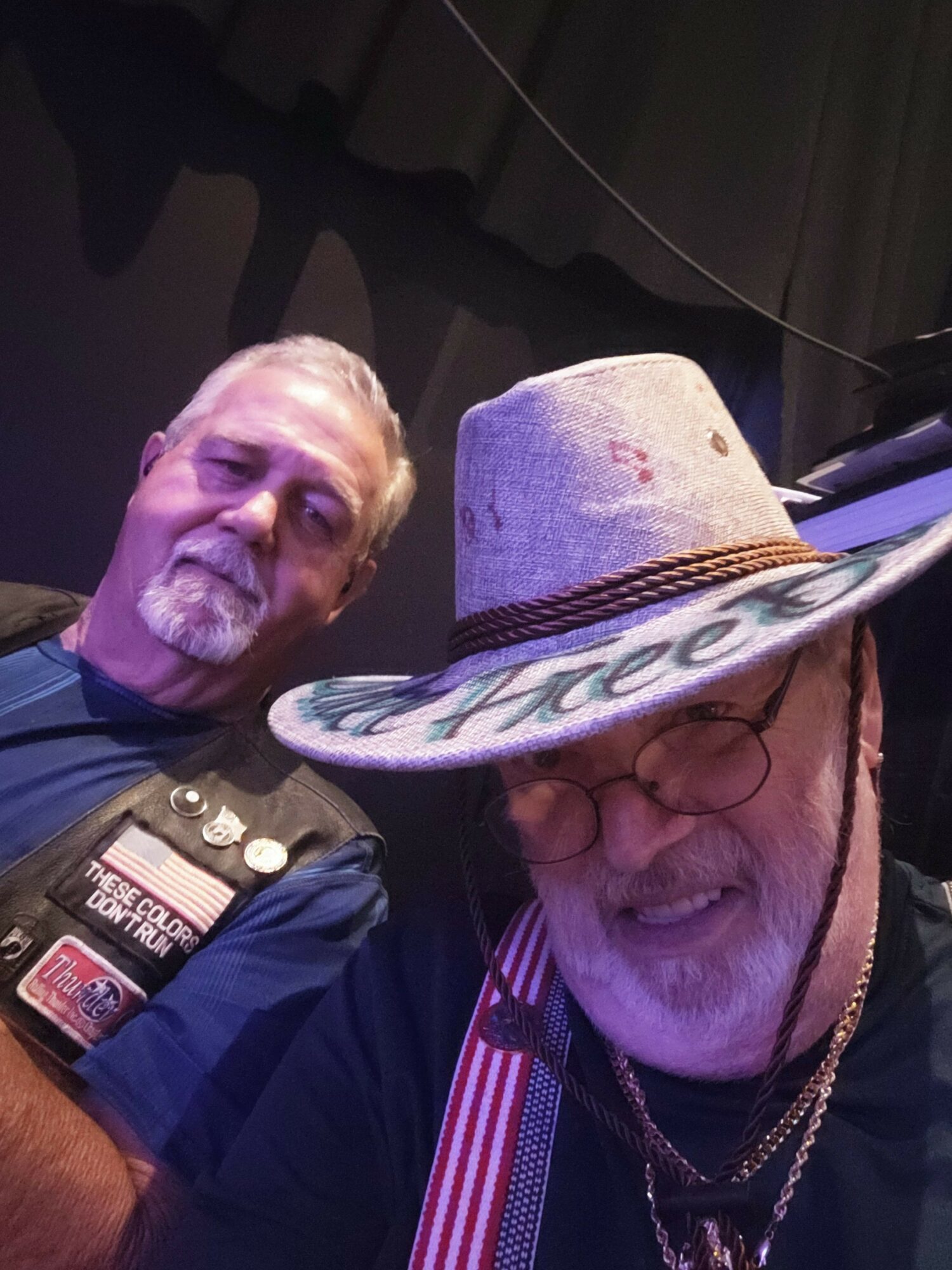 Two people, one with glasses and a hat, smiling and sitting together in a dark environment.
