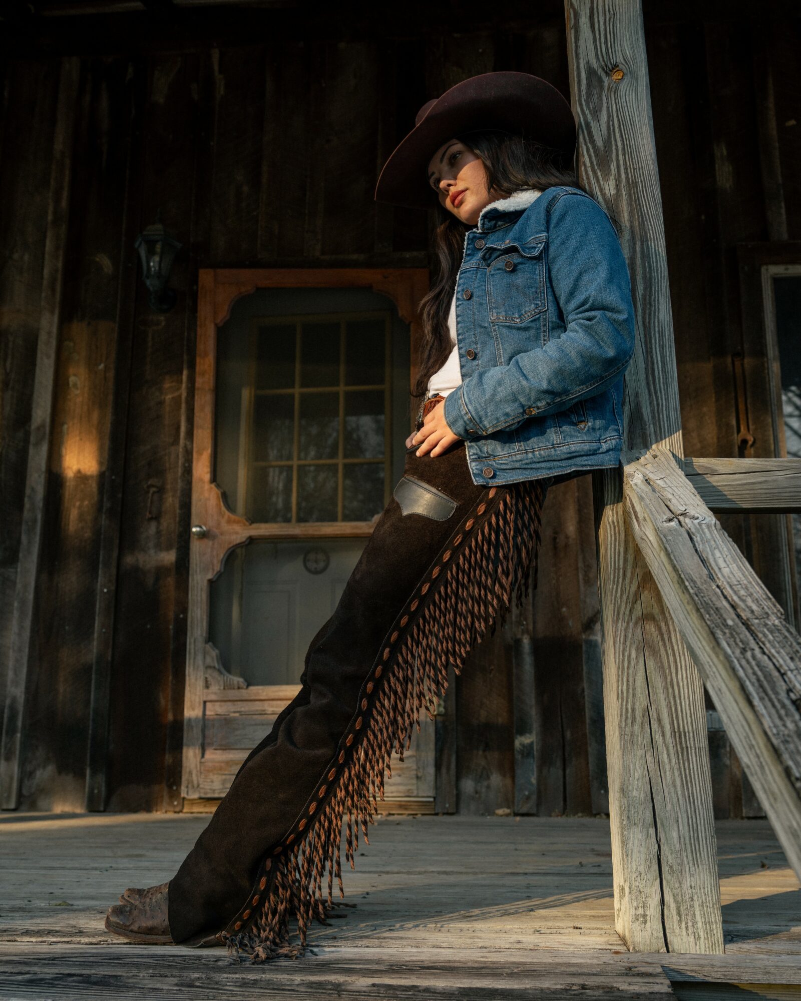 Person leaning against a wooden post, wearing a wide-brimmed hat, denim jacket, and fringed pants, standing outside a rustic building.