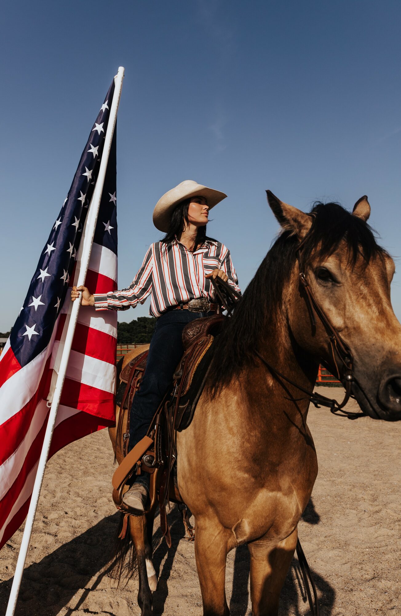 Person in cowboy hat riding a horse holding an American flag outdoors under a clear blue sky.