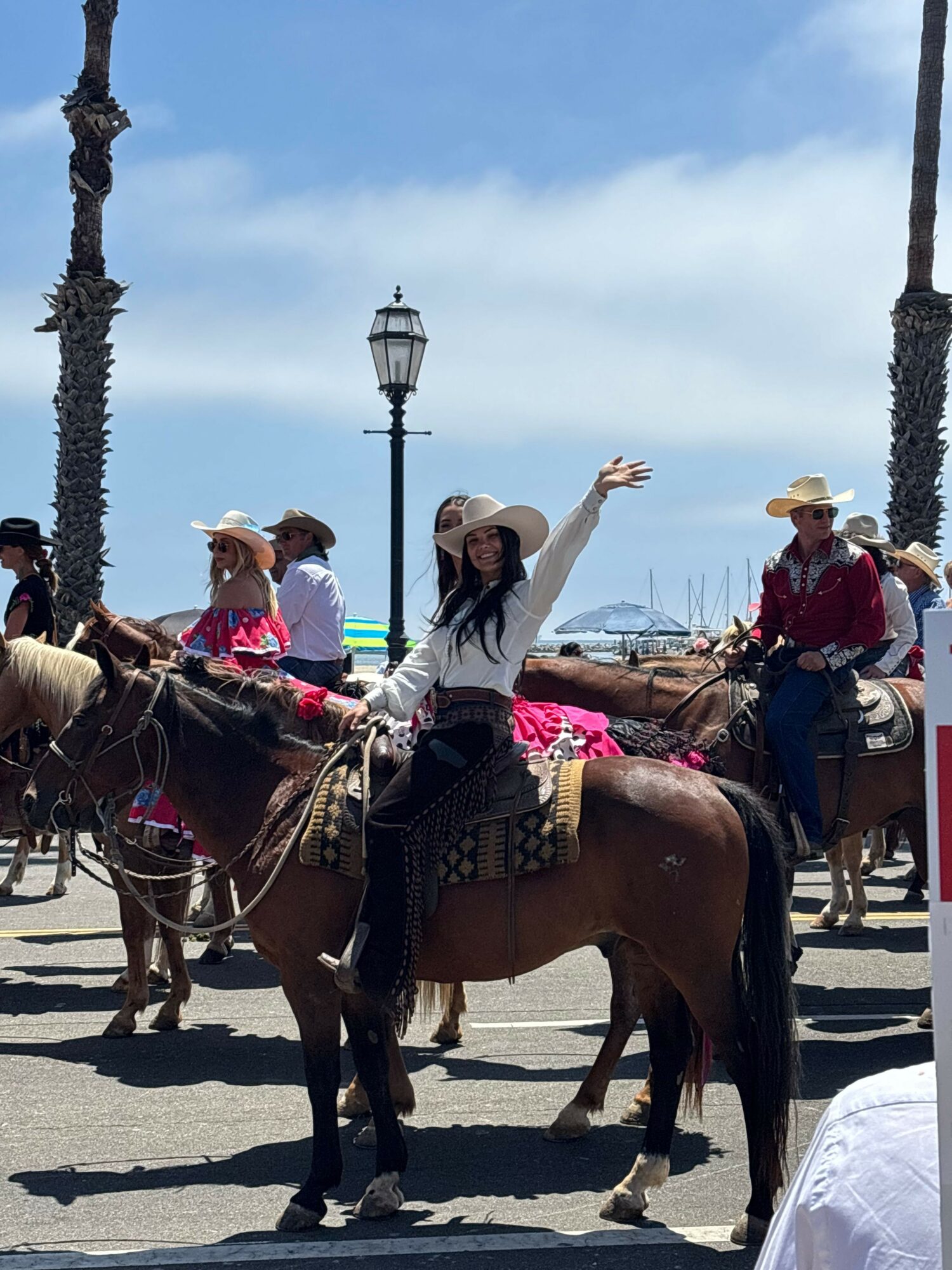 People riding horses on a street with palm trees and a lamppost in the background, some waving and smiling.