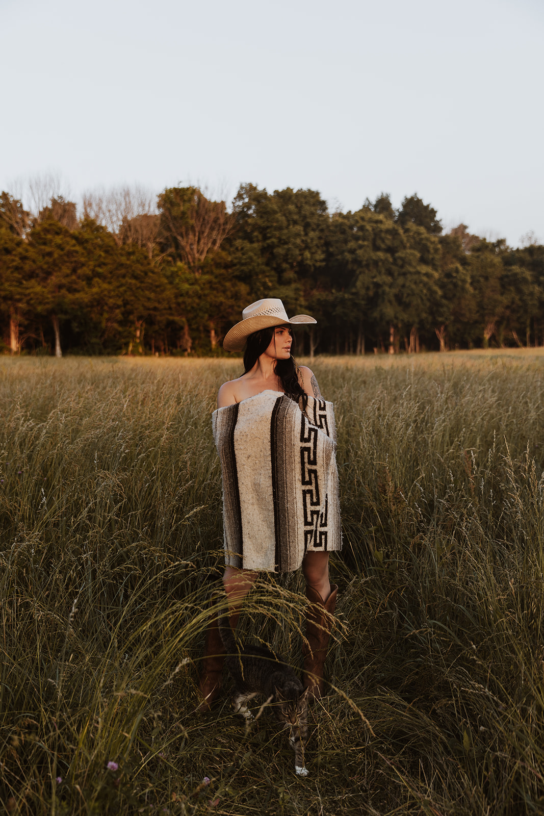 Woman in a wide-brimmed hat standing in tall grass with trees in the background during daytime.