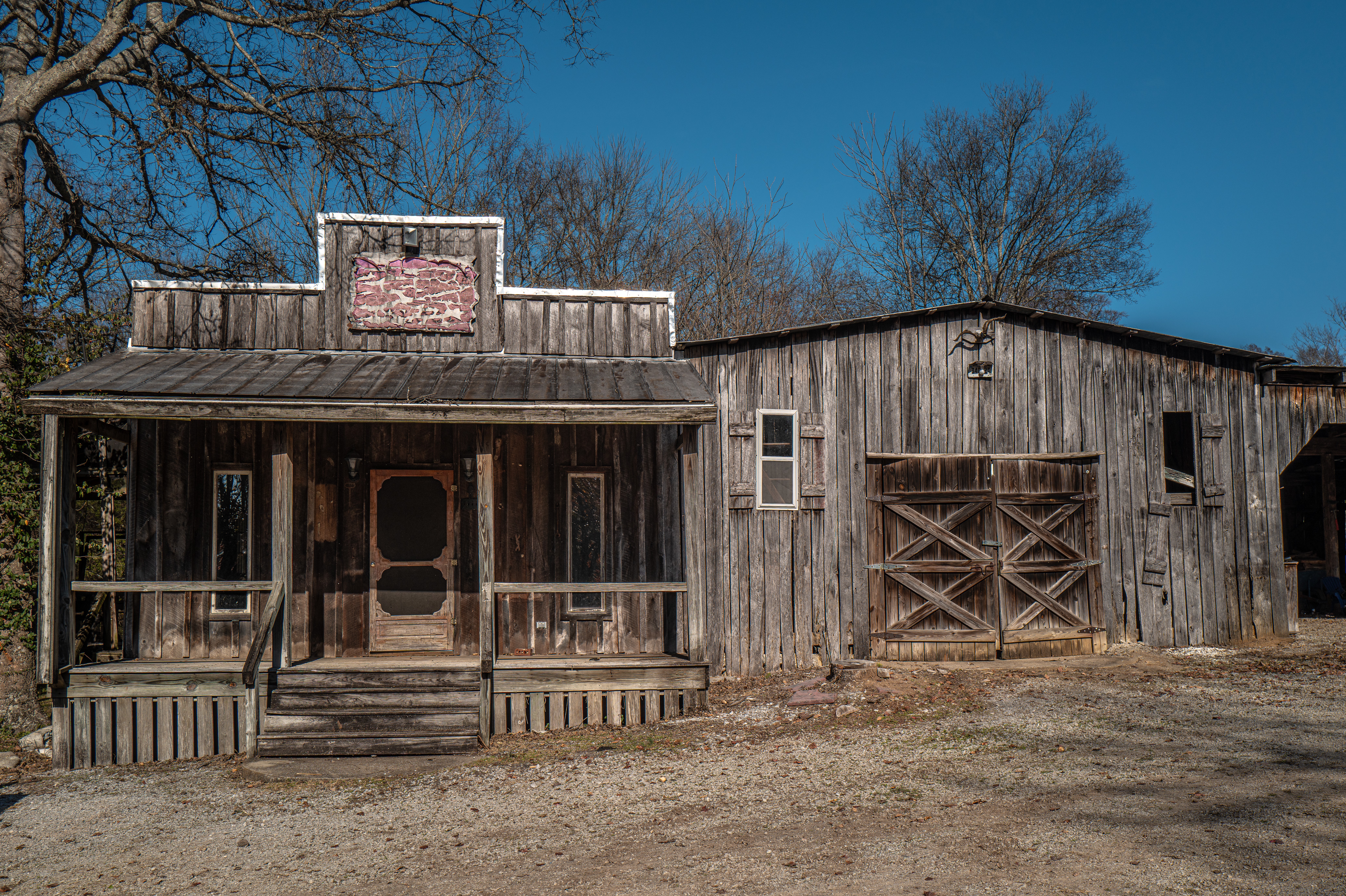 Old wooden building with a porch, stairs, and a barn door, surrounded by trees and a clear blue sky.