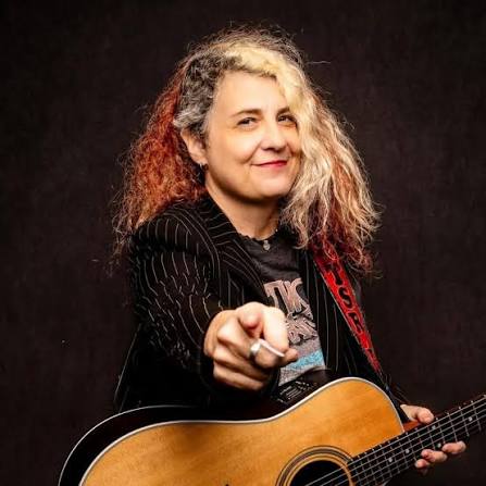 Woman with curly hair holding a guitar, pointing at the camera, smiling, dark background.