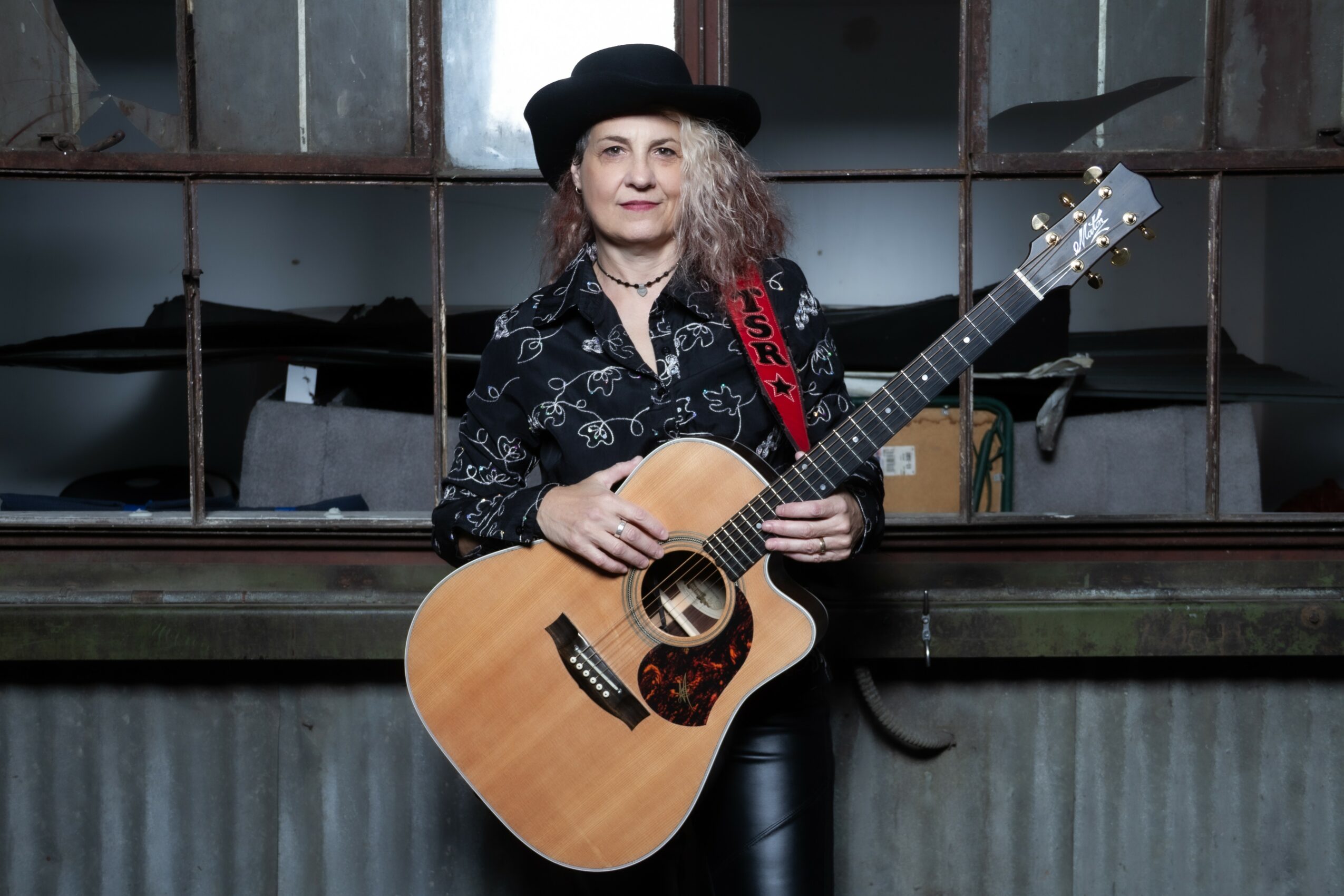 Woman with curly hair wearing a black hat and patterned shirt holding an acoustic guitar, standing in front of a window with metal bars.