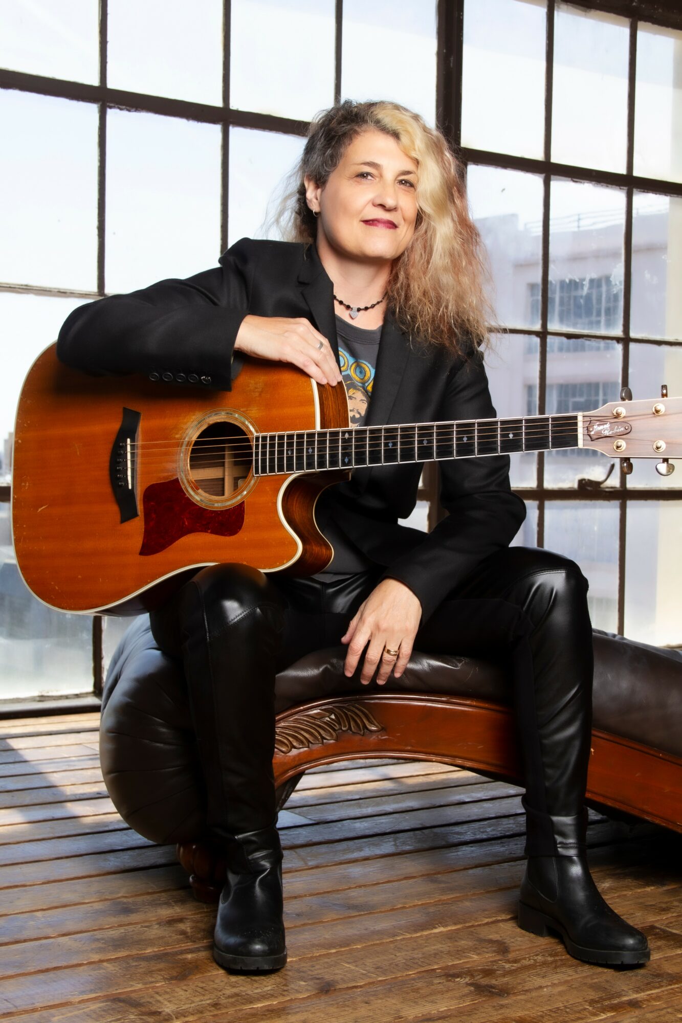 Woman with curly hair sitting on a bench holding an acoustic guitar indoors, large window behind her, cityscape view.