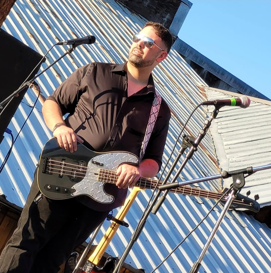 Man with sunglasses holding a bass guitar on stage outdoors, with a blue building in the background.