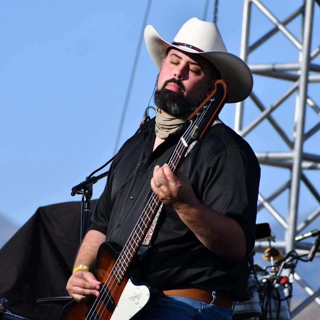 Man wearing a white cowboy hat playing an acoustic guitar on stage outdoors, with a metal structure in the background.