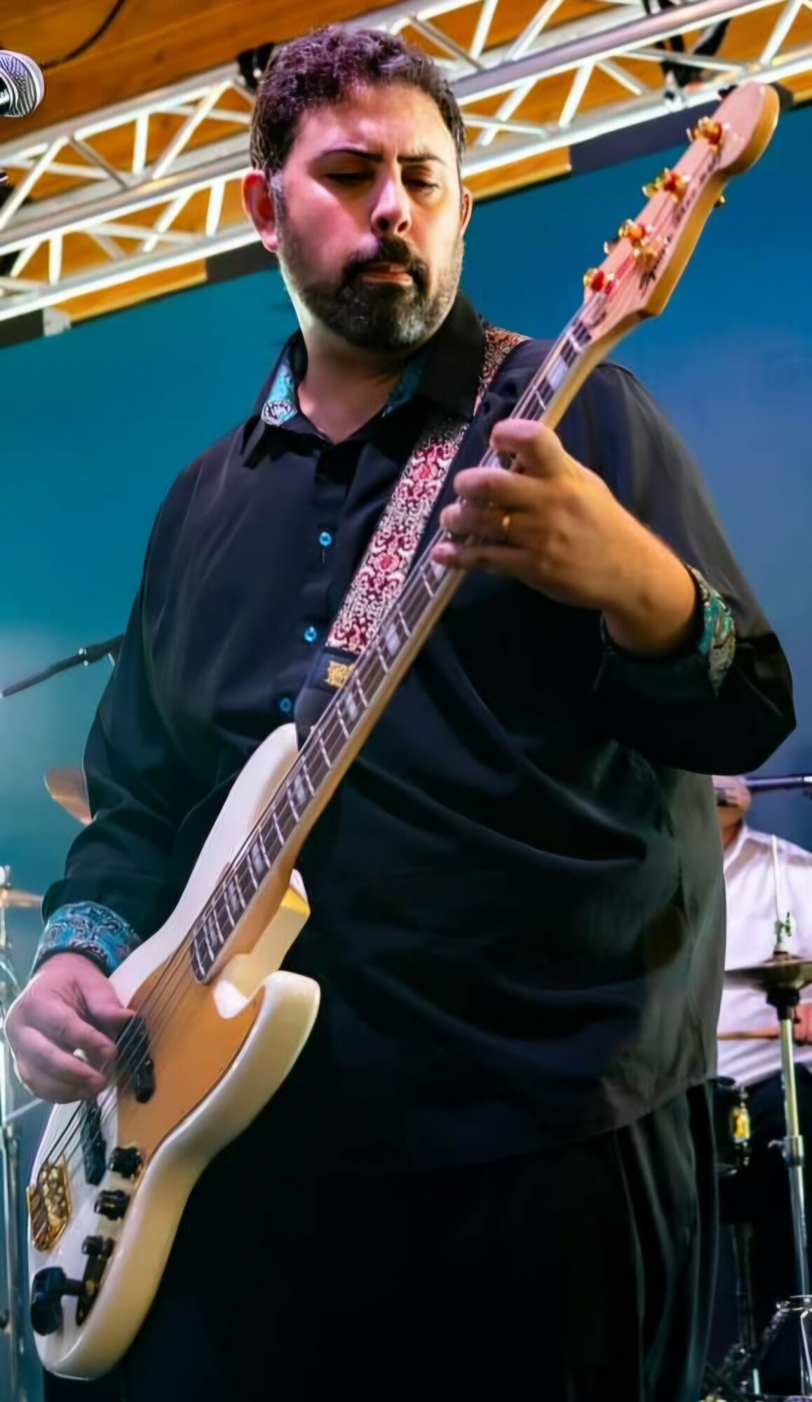 Man playing an electric guitar on stage with a yellow metal framework overhead, wearing a black shirt and looking focused.