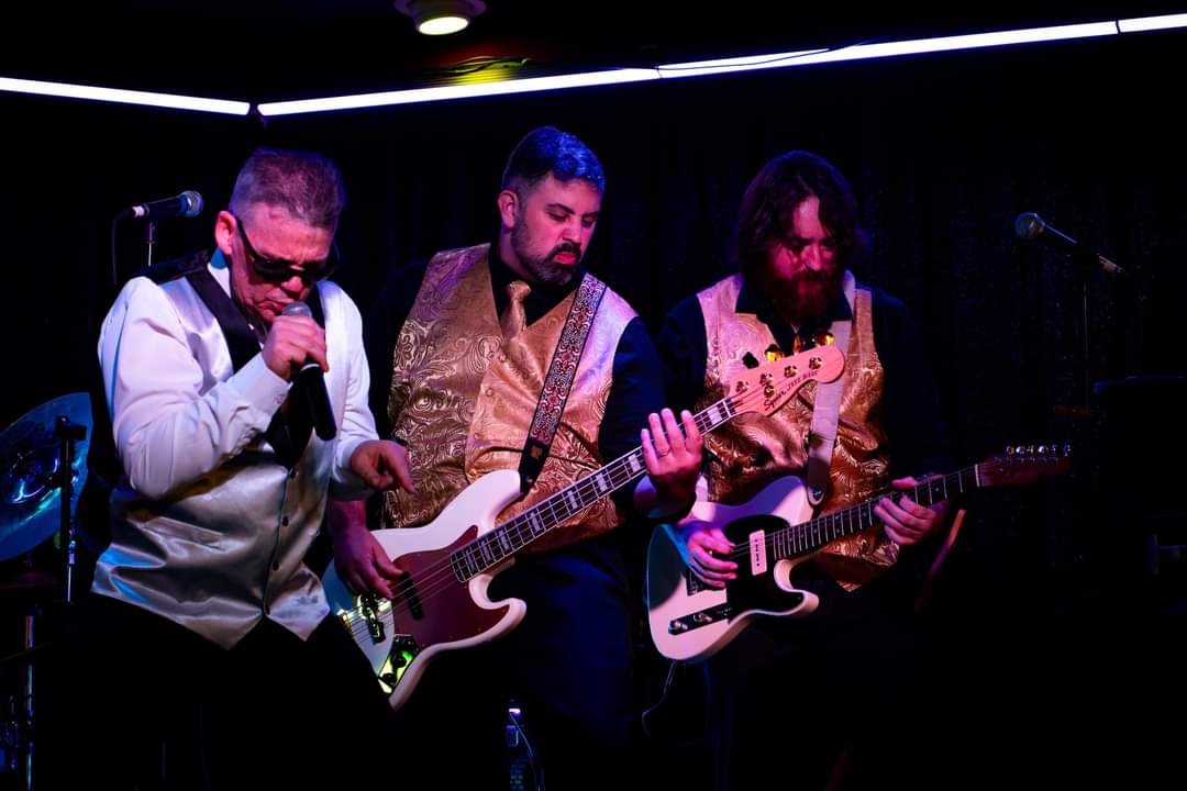 Three musicians perform on stage with guitars and microphone, dark background, stage lighting, and black curtains.