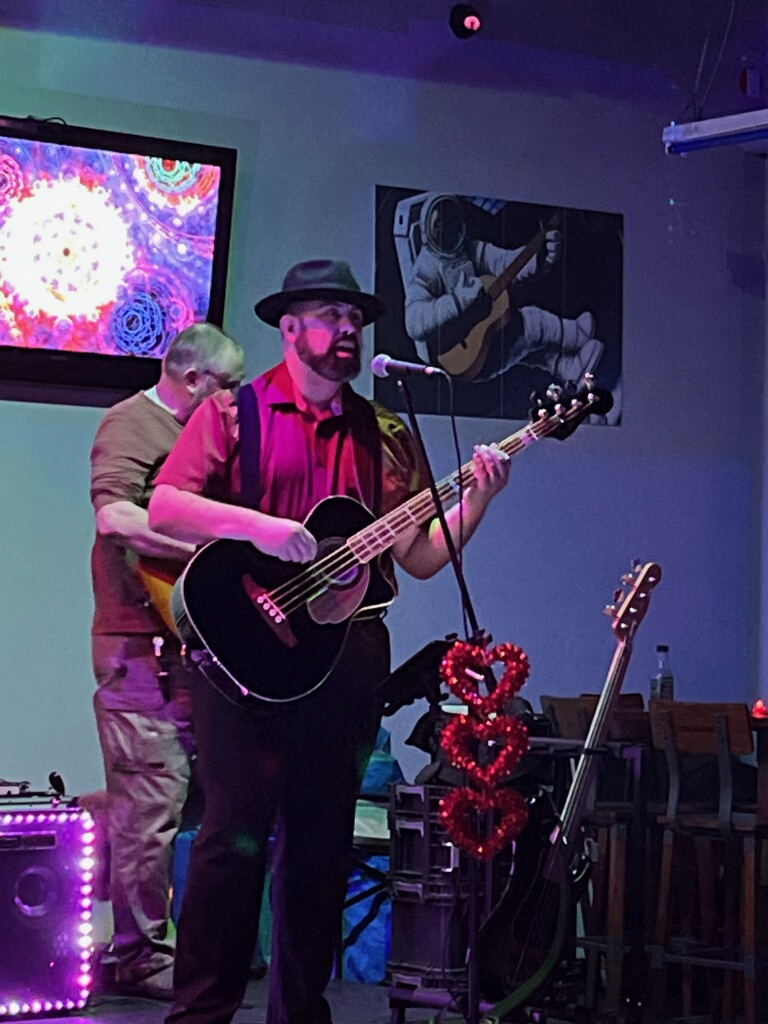 Man with a beard wearing a hat and red shirt playing an acoustic guitar on stage, with a colorful screen and artwork in background.