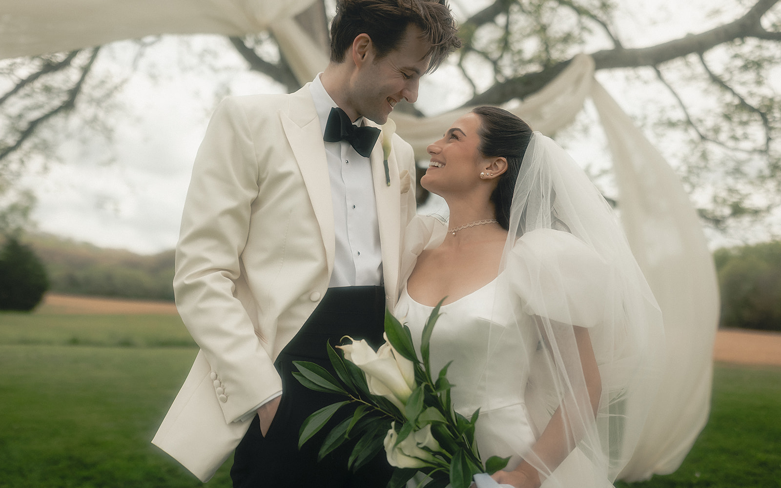 A bride and groom in wedding attire smiling at each other outdoors, holding a bouquet of white flowers.