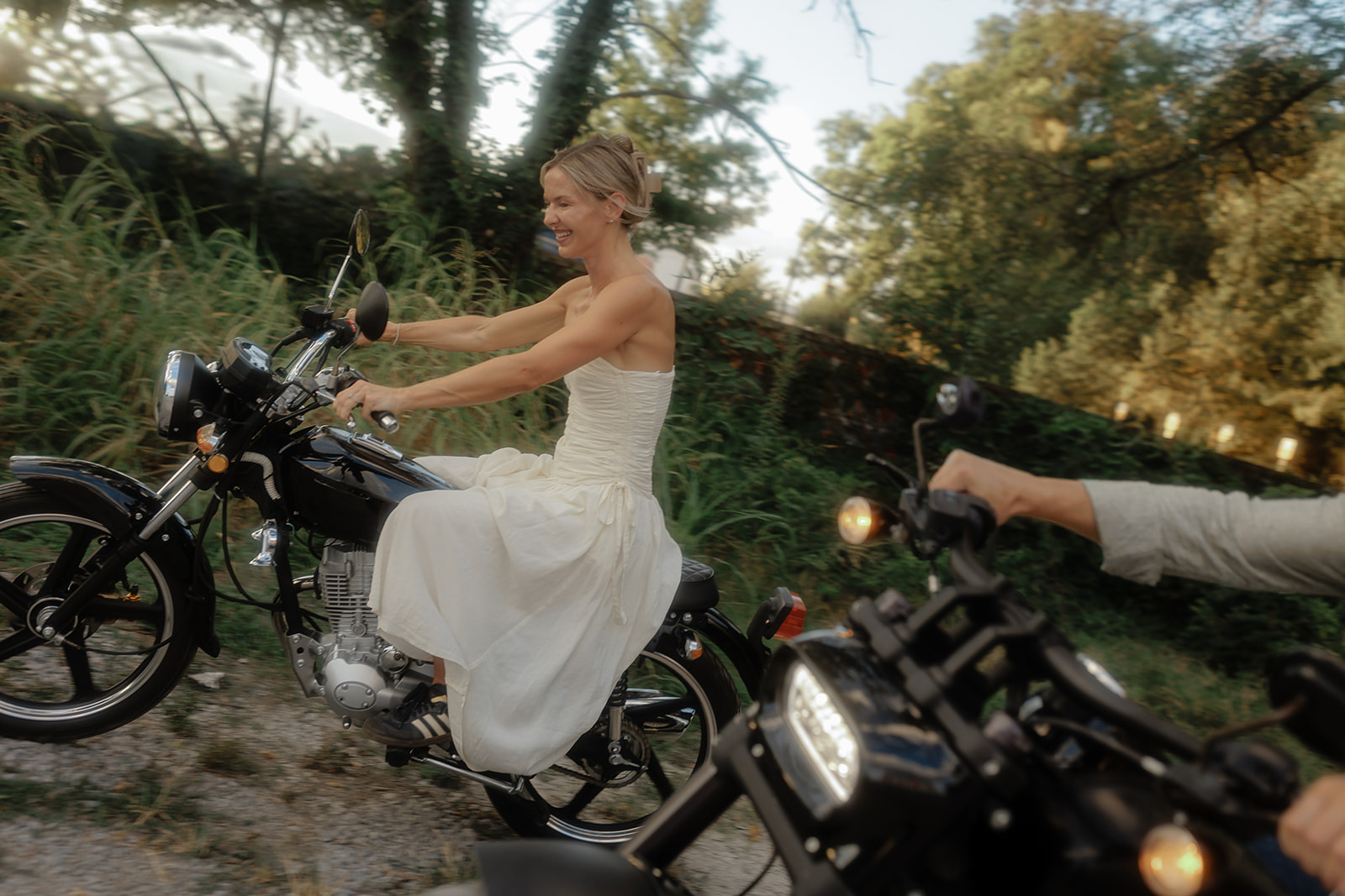 Woman in a white dress riding a motorcycle outdoors with trees in the background.