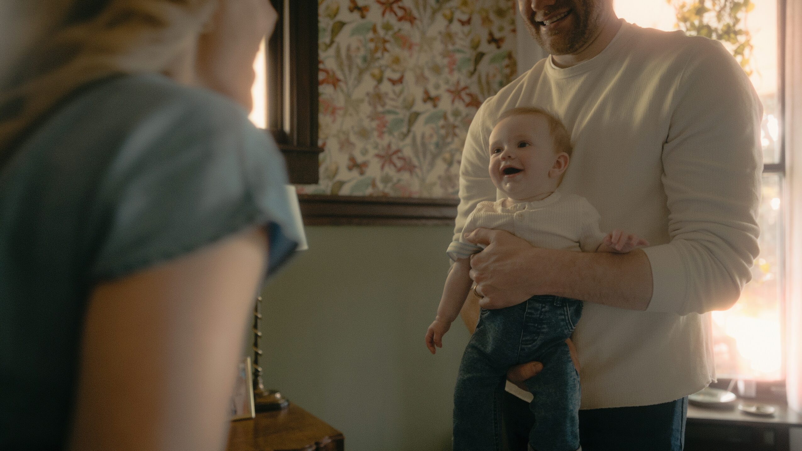 Person holding a smiling baby, with another person partially visible in the foreground, in a room with floral wallpaper and sunlight.