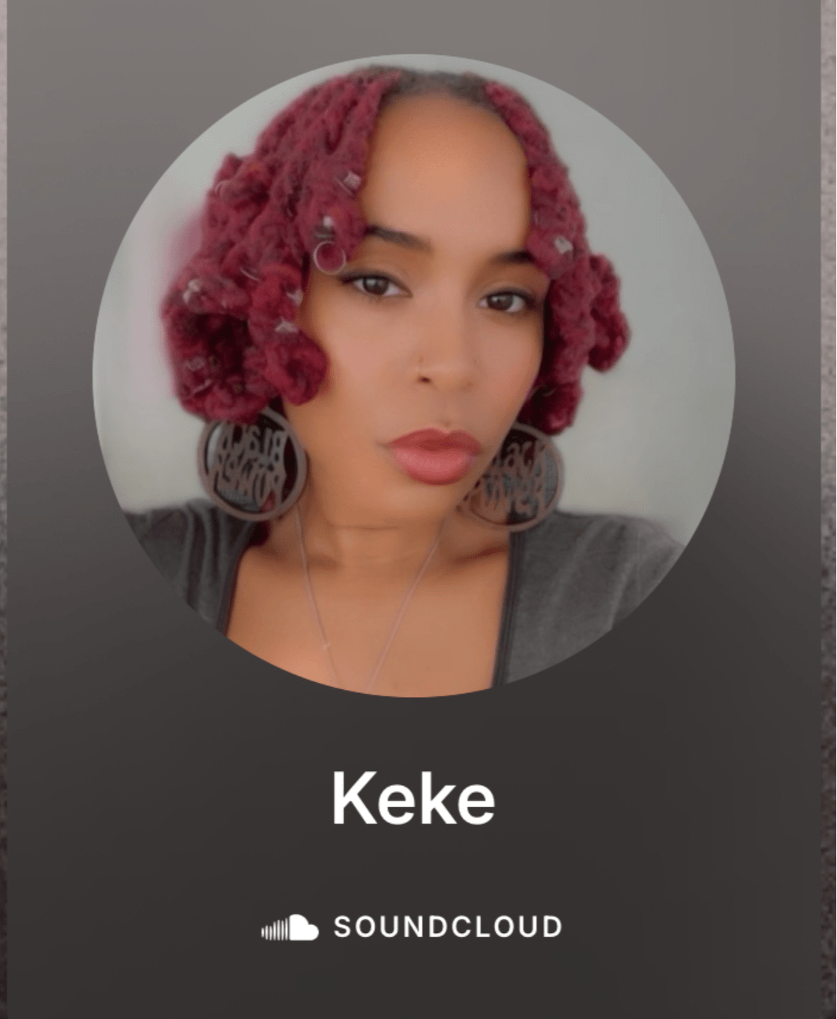 Portrait of a woman with pink curly hair, wearing large earrings and a dark top, looking at the camera.
