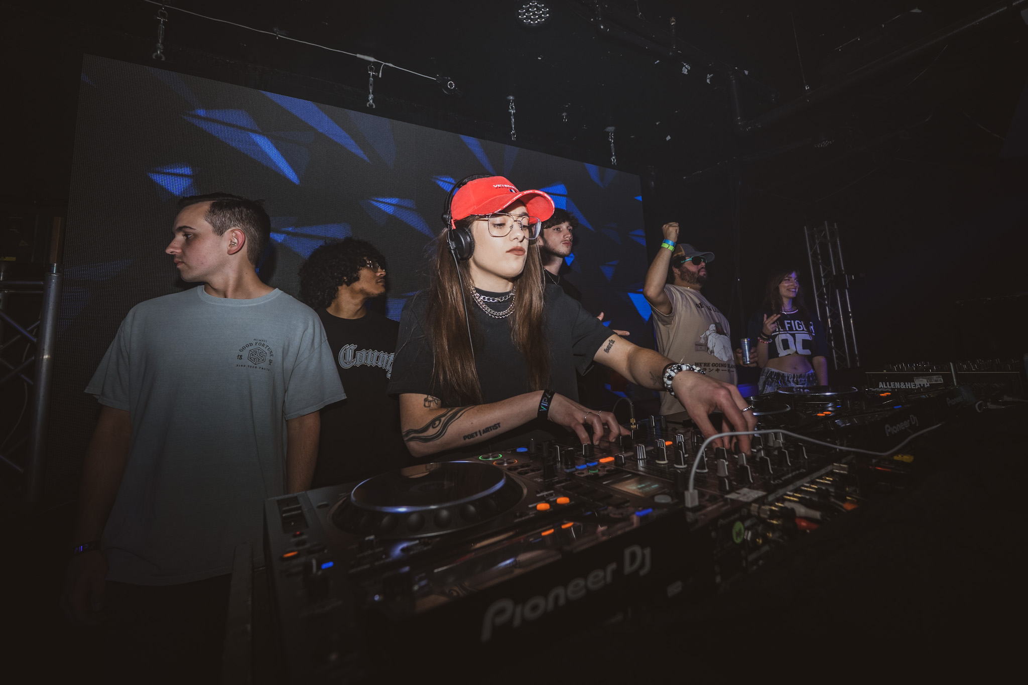 DJ wearing a red cap and black shirt operates a Pioneer DJ mixer at a club with people in the background.