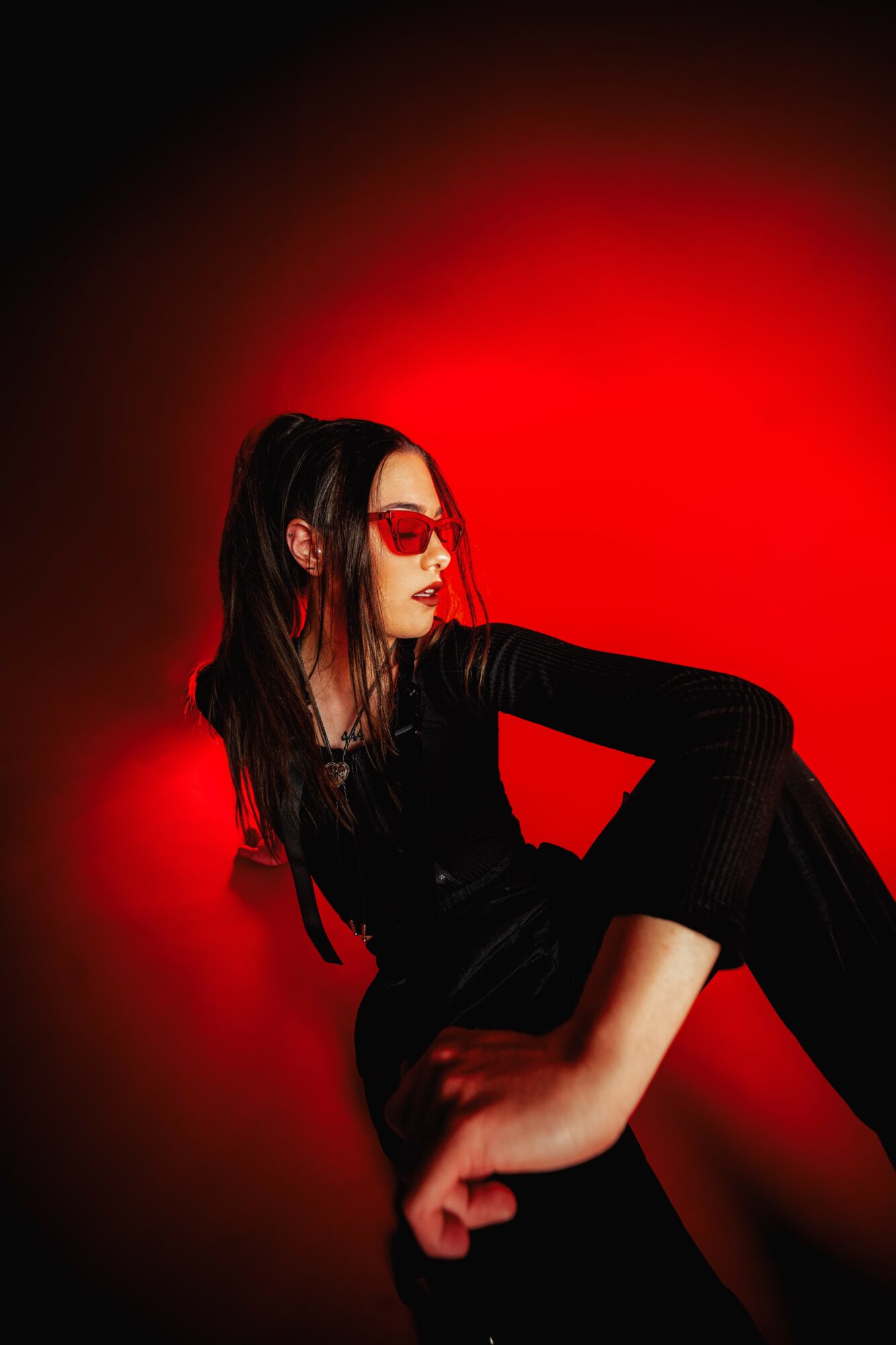 Woman with long dark hair wearing red sunglasses and black outfit, posed against a red and black background.