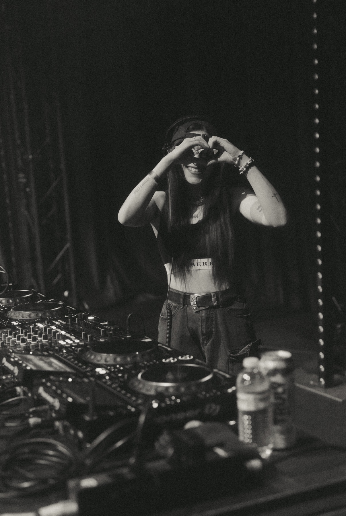 Person with long hair taking a photo in a mirror, standing behind DJ equipment, in a dark setting.
