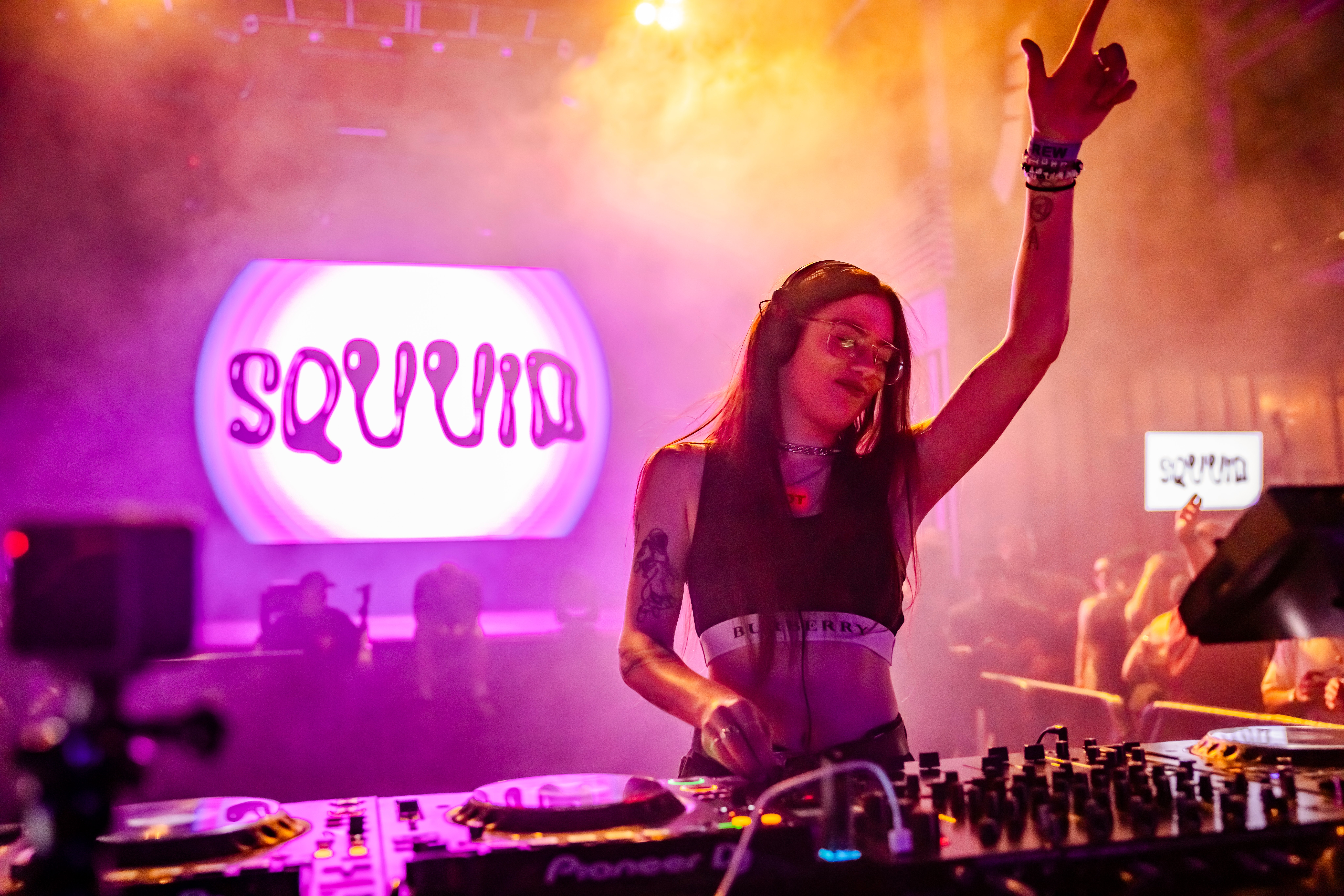 Female DJ with long hair and tattoos raises her hand in a club with colorful lights and a large 'SQUIN' sign in the background.