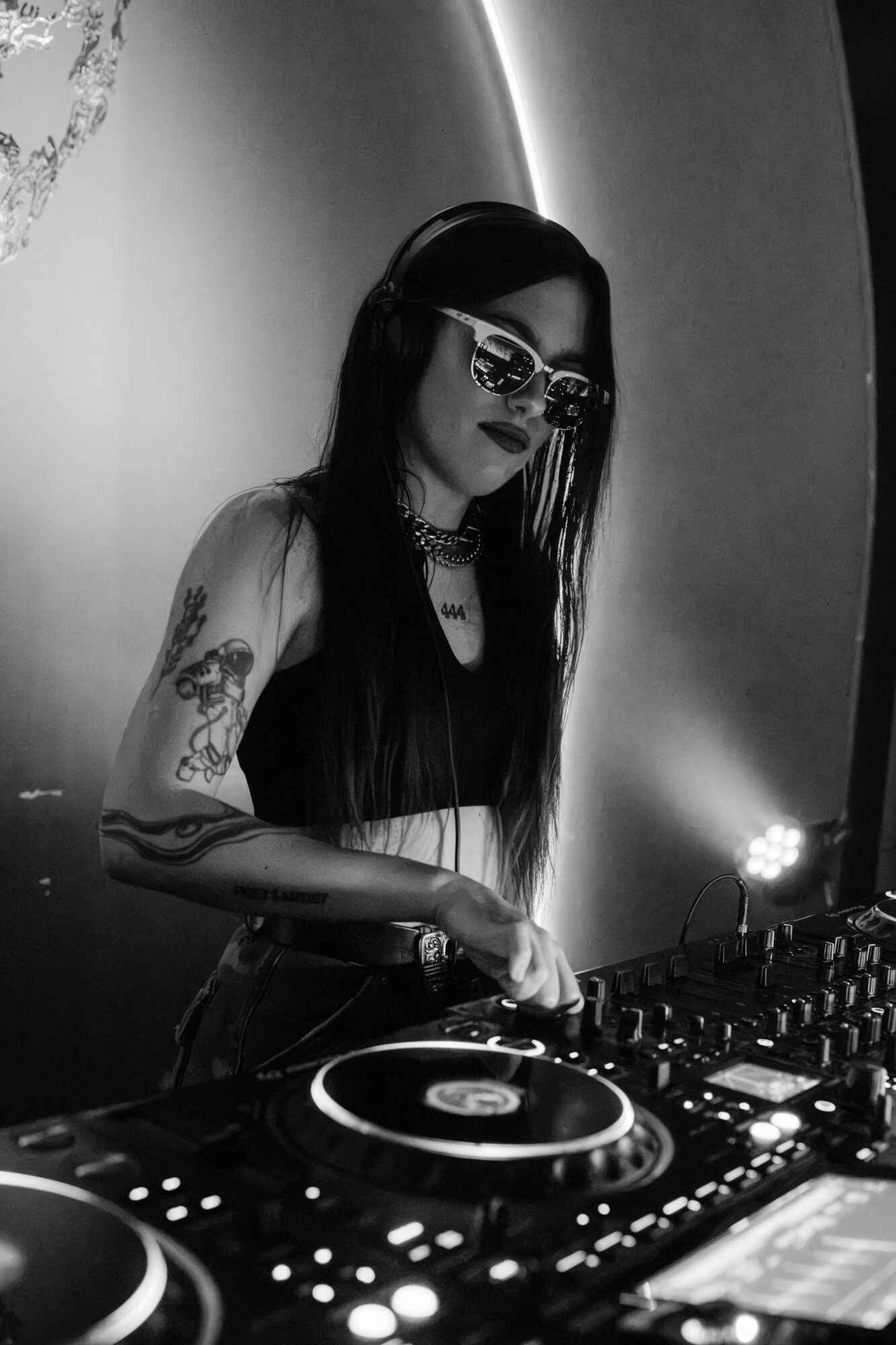 Woman with long dark hair, sunglasses, tattoos, and headphones DJing with equipment in a dimly lit setting.