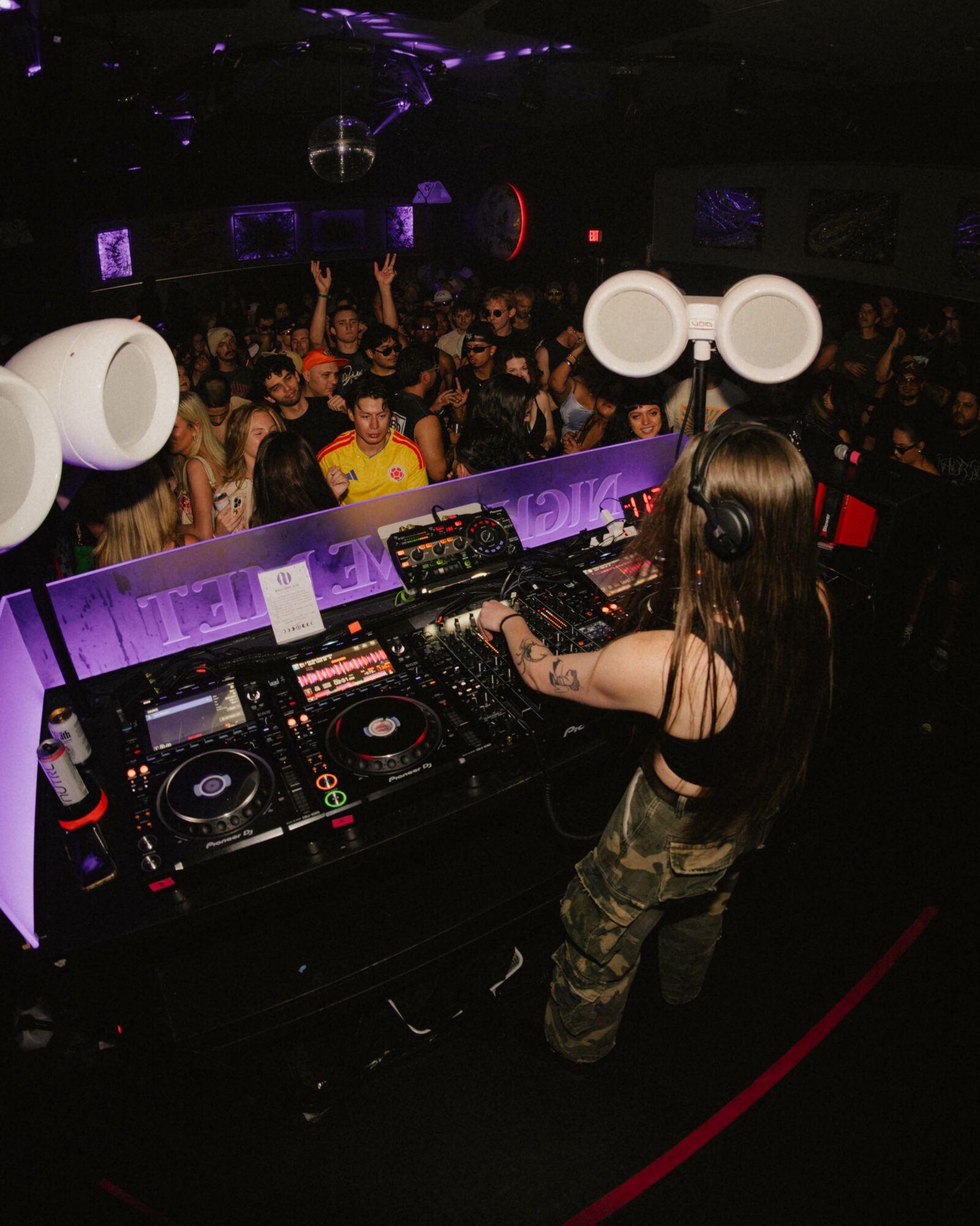 DJ woman with headphones operating turntables in a nightclub with a crowd dancing, purple lighting, and speakers, viewed from above.