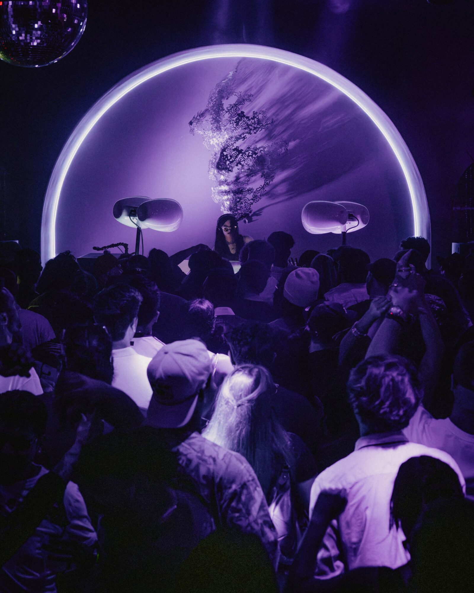 Crowd watching a DJ perform on stage with a circular light display and purple lighting, in a nightclub or concert setting.