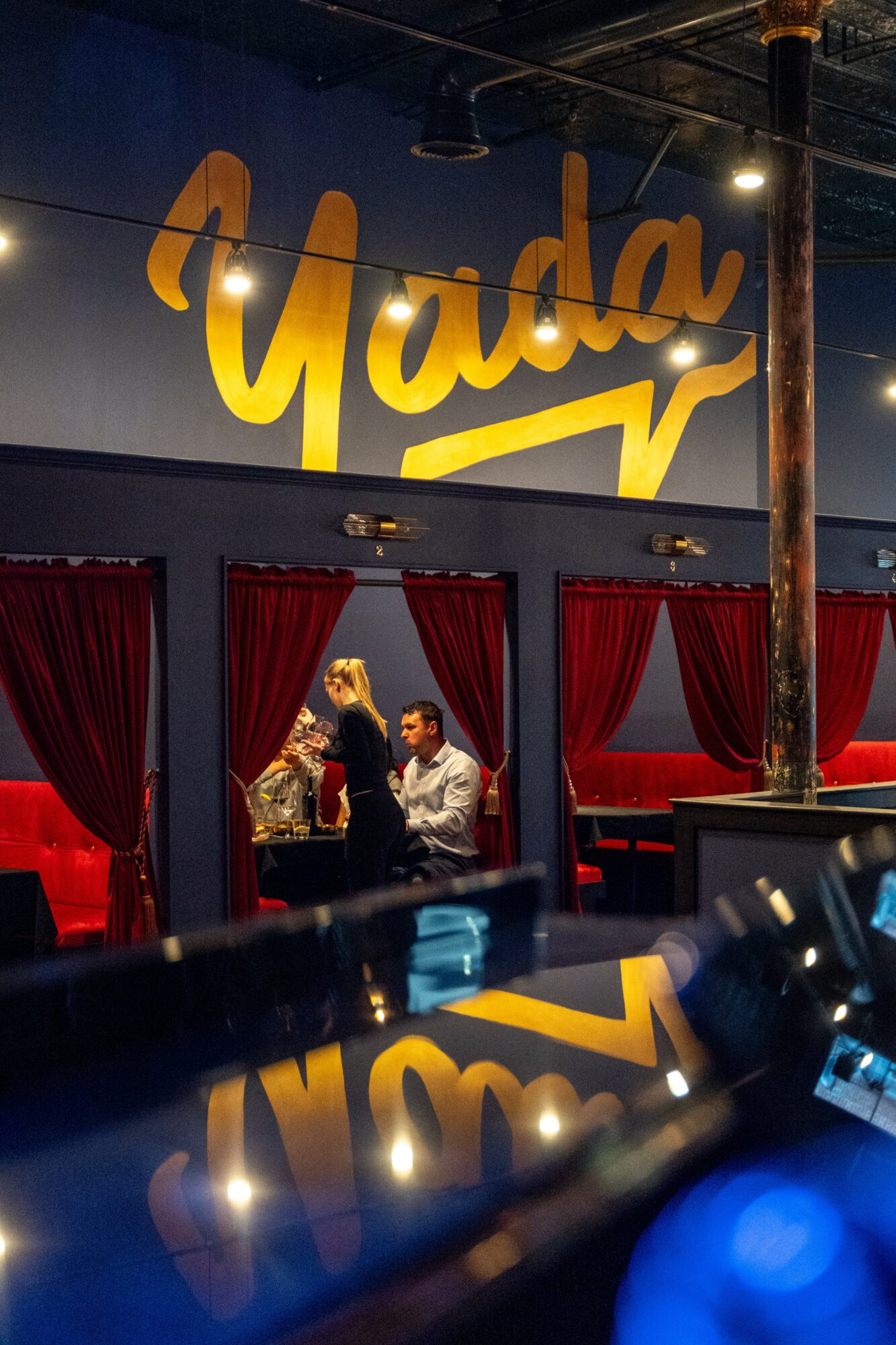People sitting at a bar with red curtains and a large yellow sign reading 'Yada' on the wall.