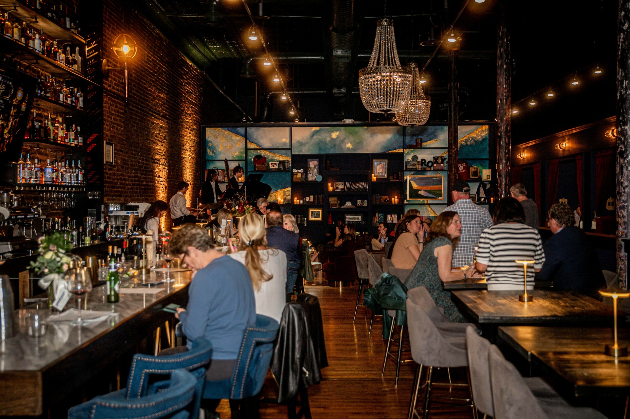 People sit at tables and a bar in a dimly lit restaurant or bar with brick walls and a chandelier, large screens on the wall.