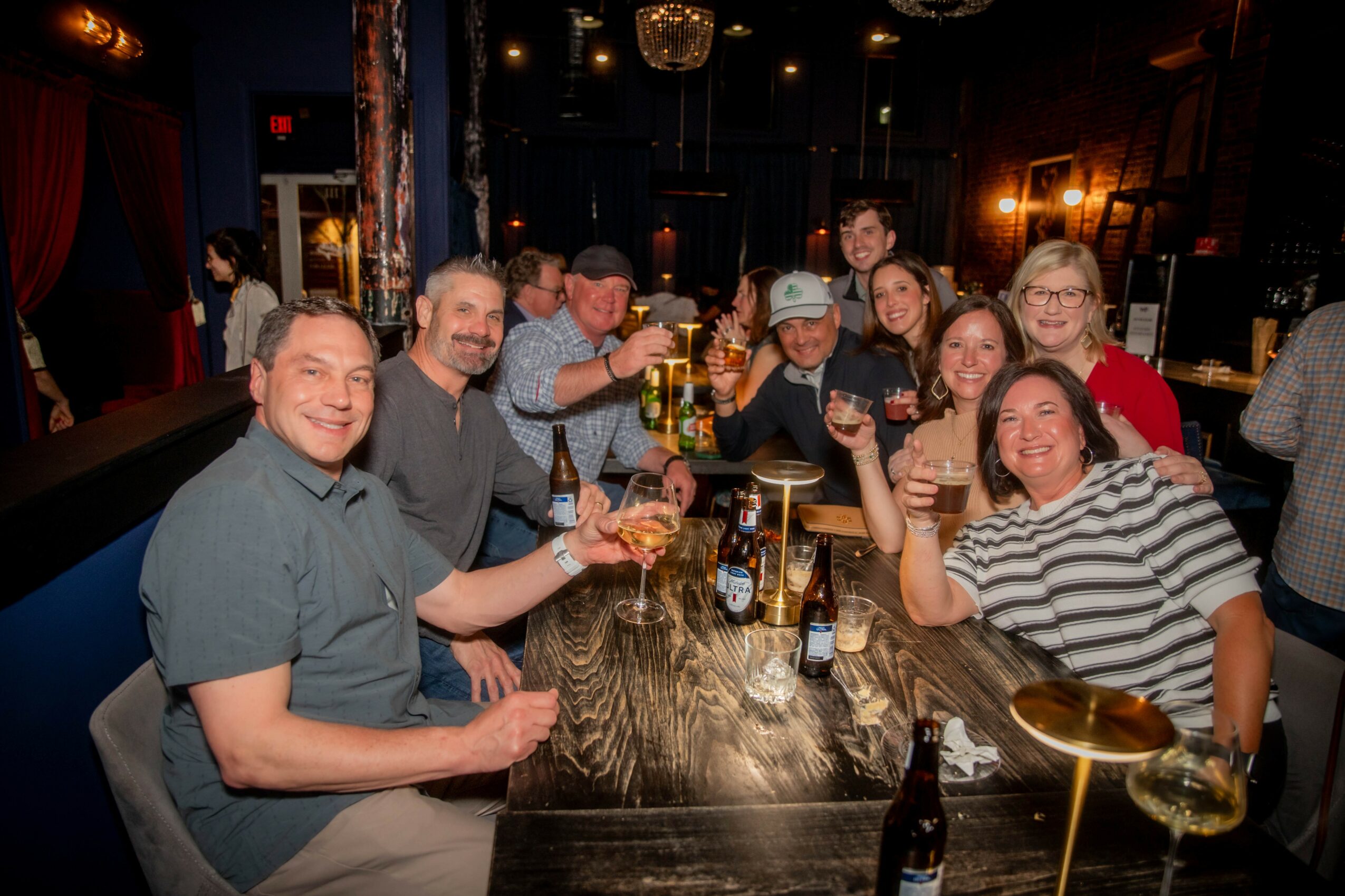 Group of people sitting at a bar table, smiling, holding drinks, celebrating together, in a dimly lit venue.