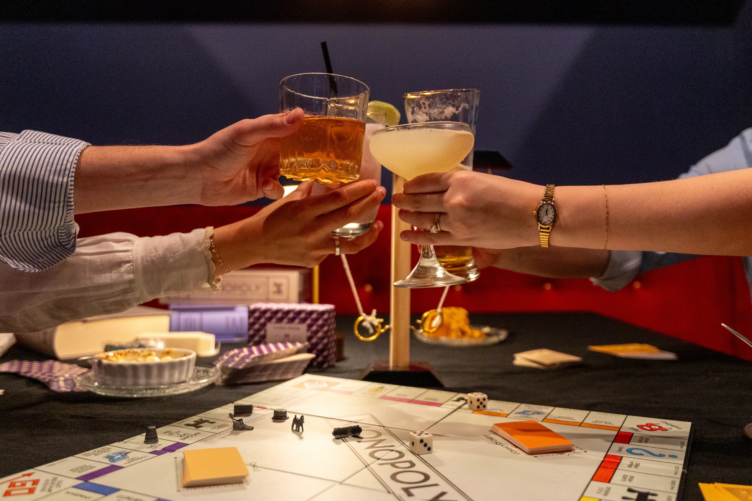 Two people clink glasses with drinks over a table, with game pieces and a board visible, in a social setting.