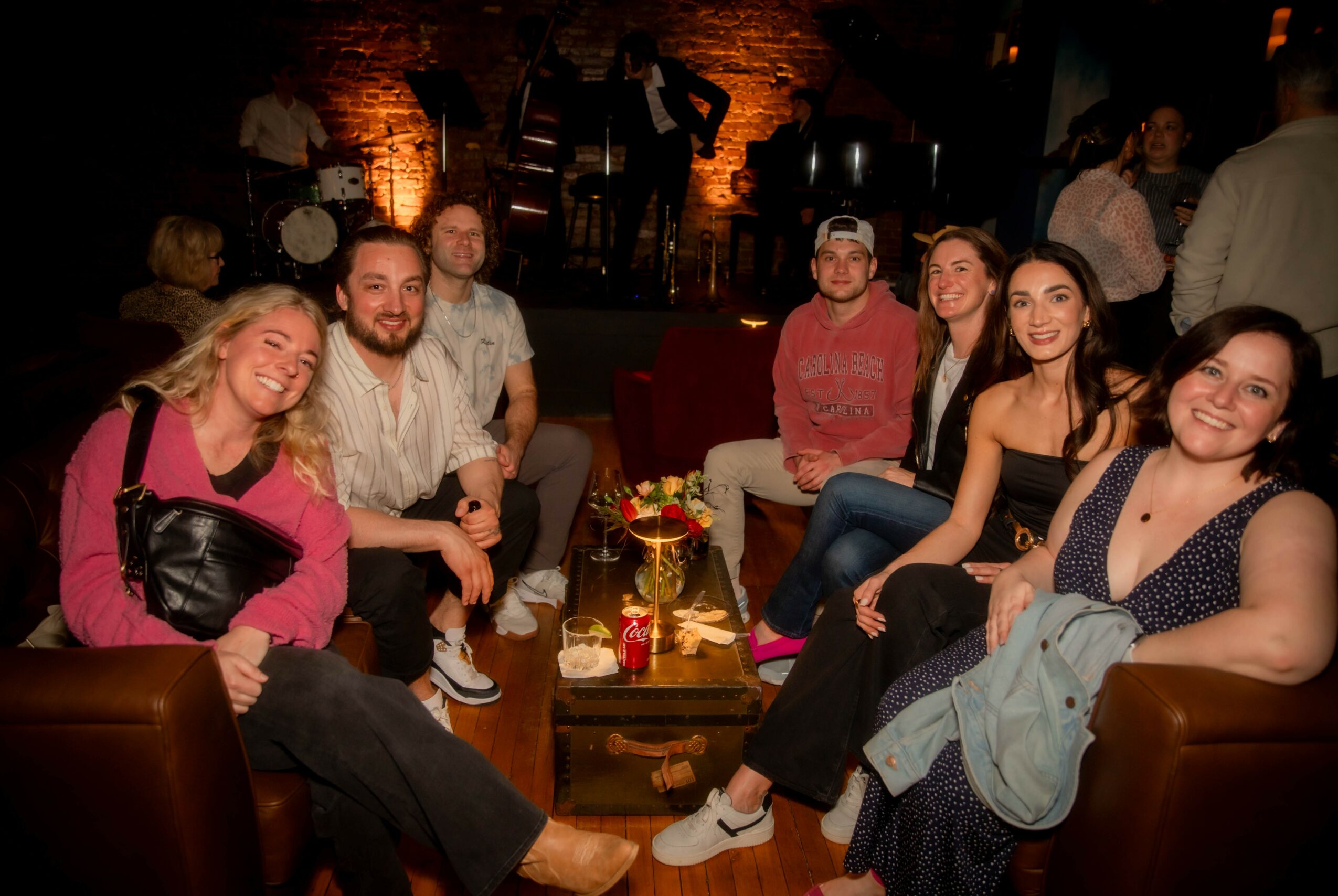 Group of seven people sitting on couches in a dimly lit bar or lounge, smiling and enjoying each other's company.