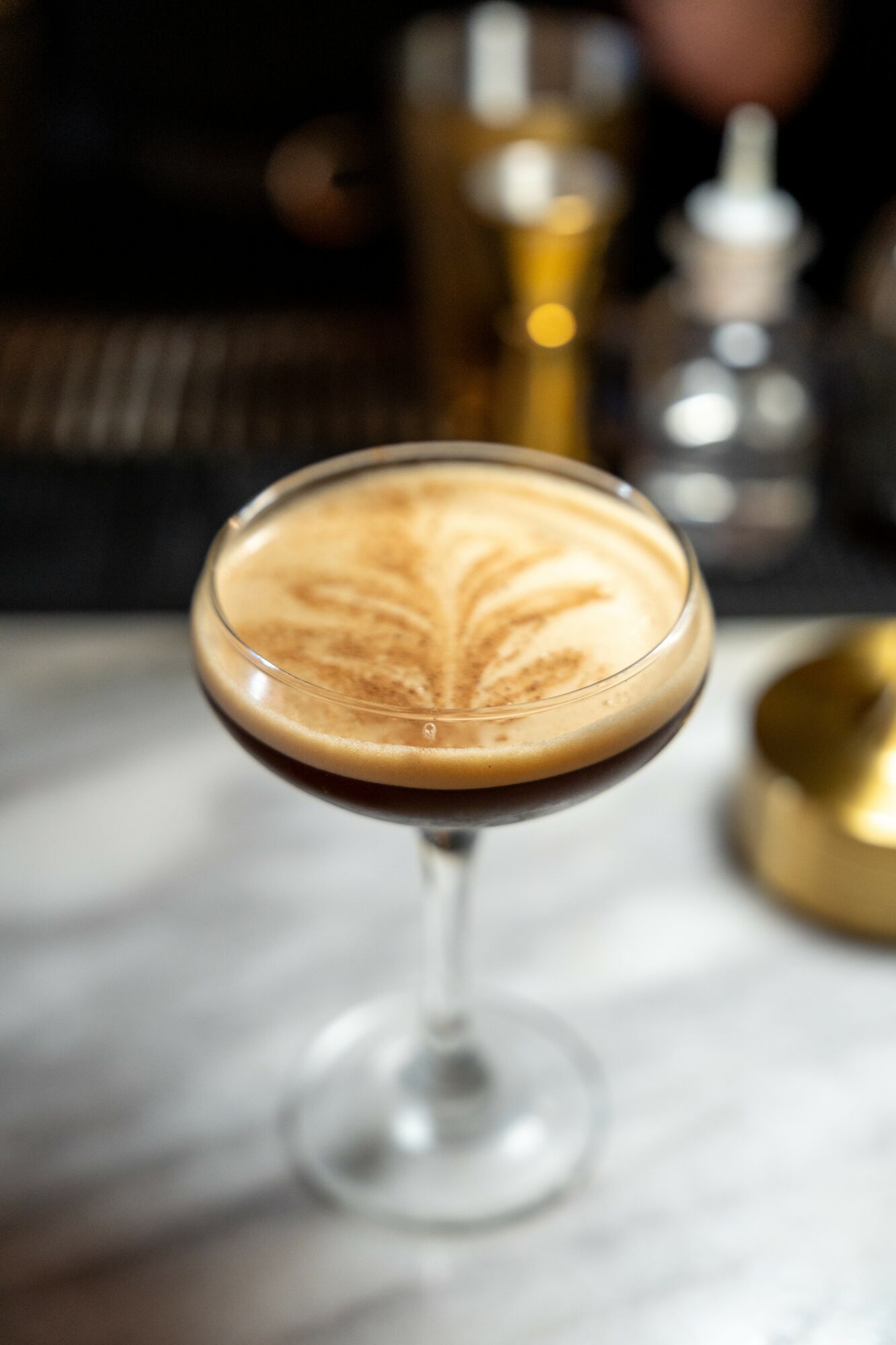 A cocktail glass with a frothy drink and latte art on top, placed on a white surface with blurred background.